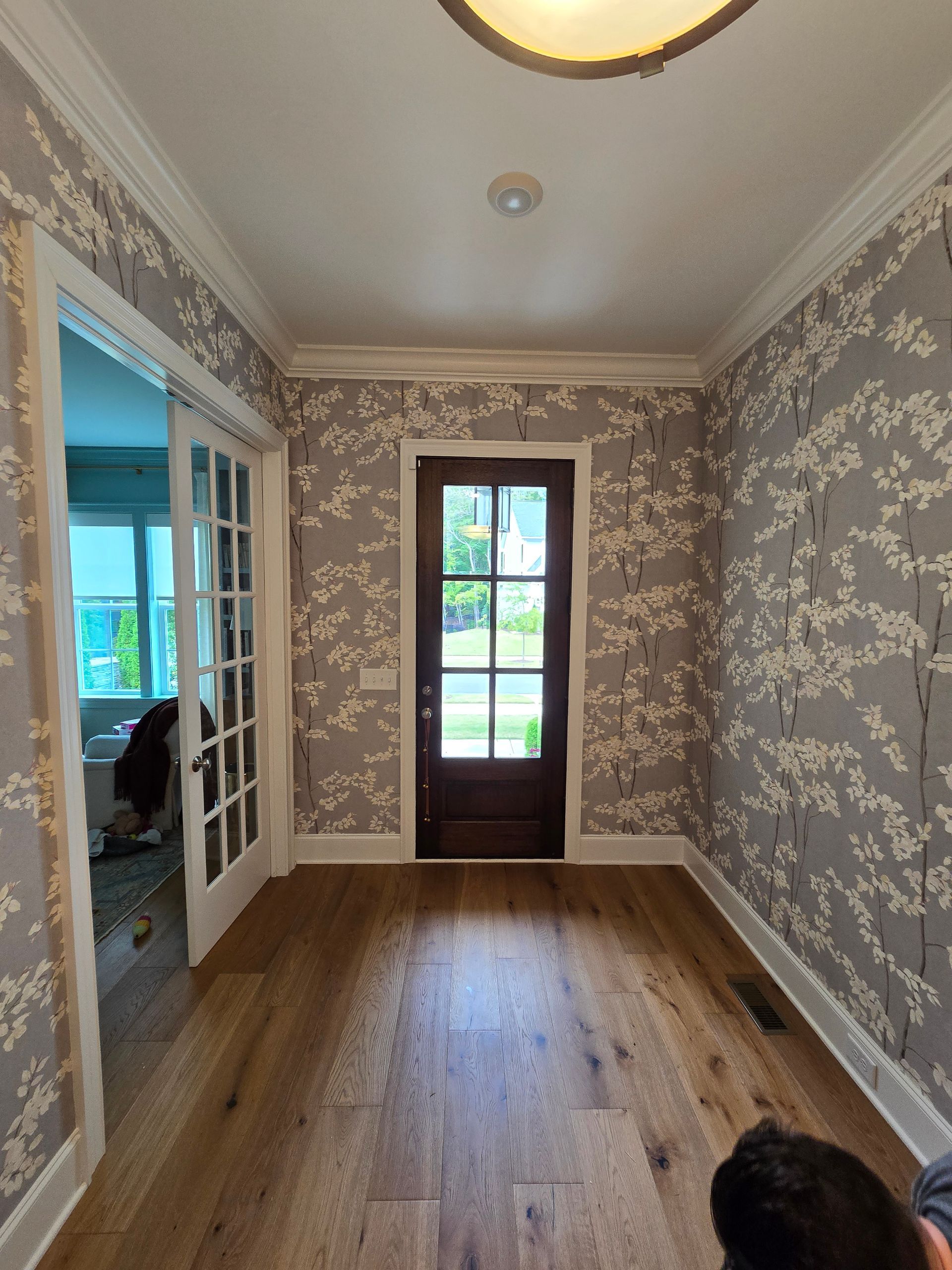 A foyer with floral gray wallpaper, hardwood floors, a dark wood front door, and a glass-paneled double door entrance.