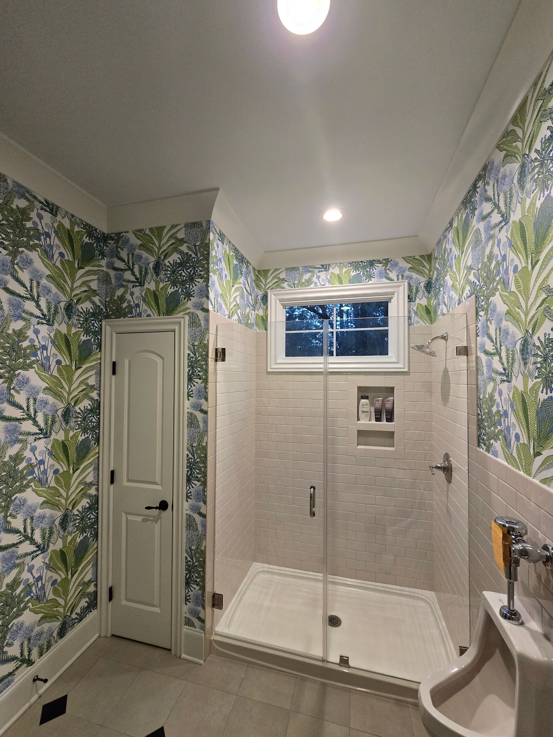 Bathroom with a glass-enclosed shower, cream tiled walls, a white door, and vibrant blue and green botanical wallpaper.