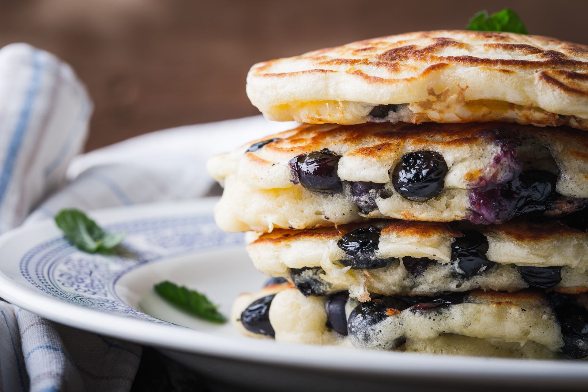 Stack of blueberry pancakes on a white plate, with mint leaves, brown background.