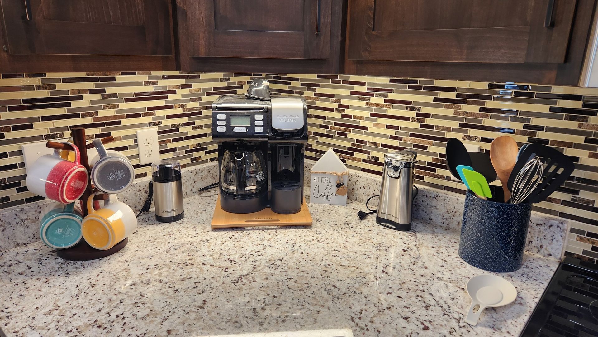 Coffee station with coffee maker, mugs, utensils, and a patterned backsplash.