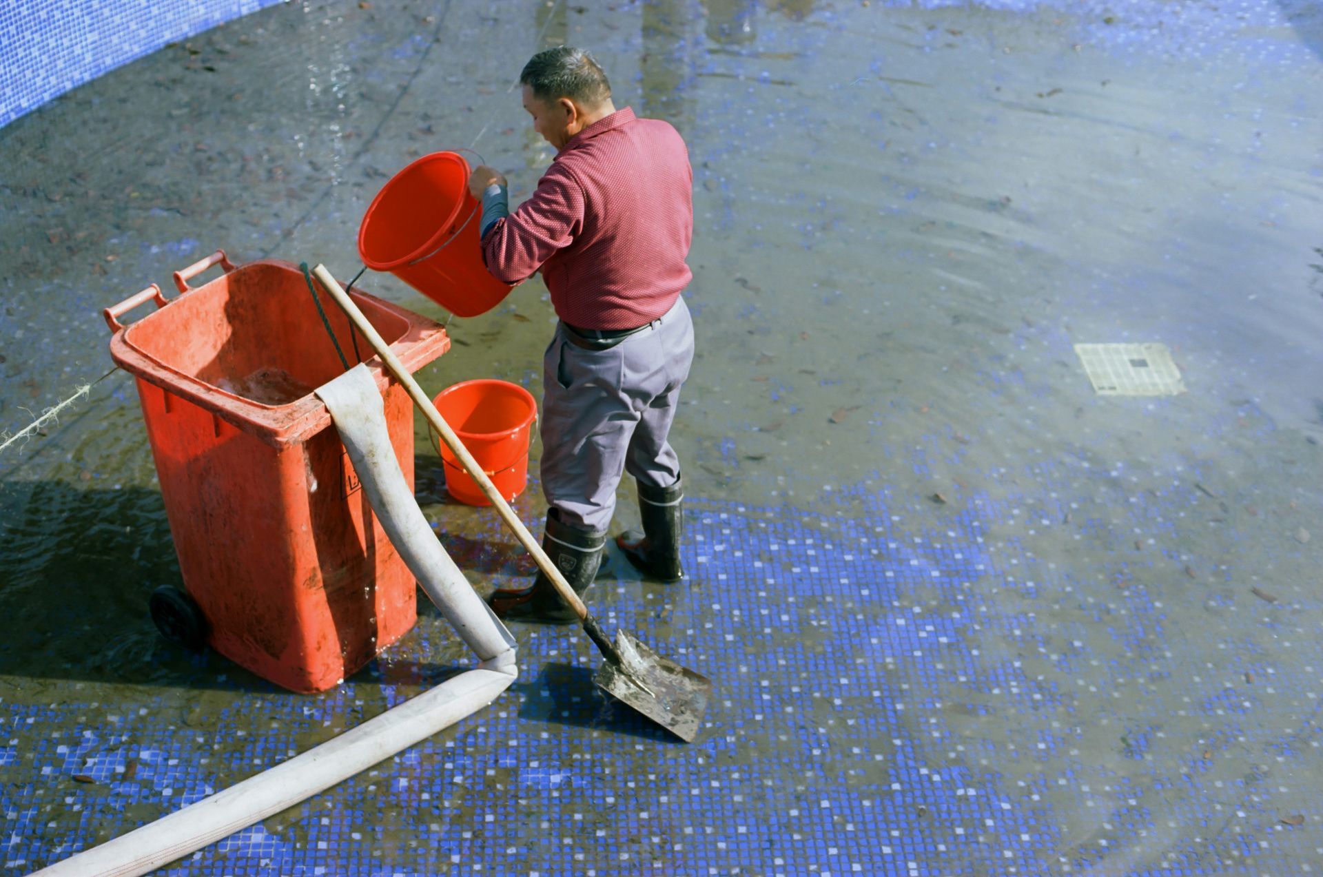 Worker pouring material into orange bins beside a vacuum hose on a blue-speckled concrete floor