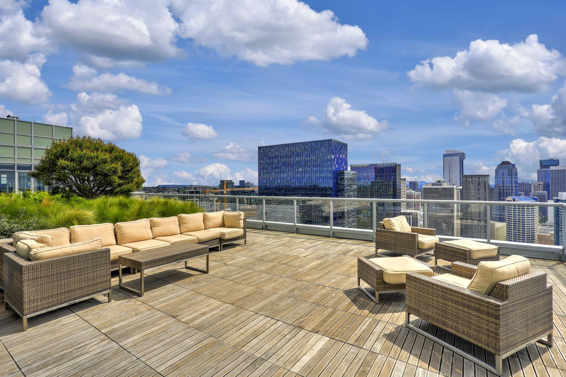 A rooftop patio with wicker lounge furniture on wooden tiles, overlooking a city skyline under a blue sky with clouds.