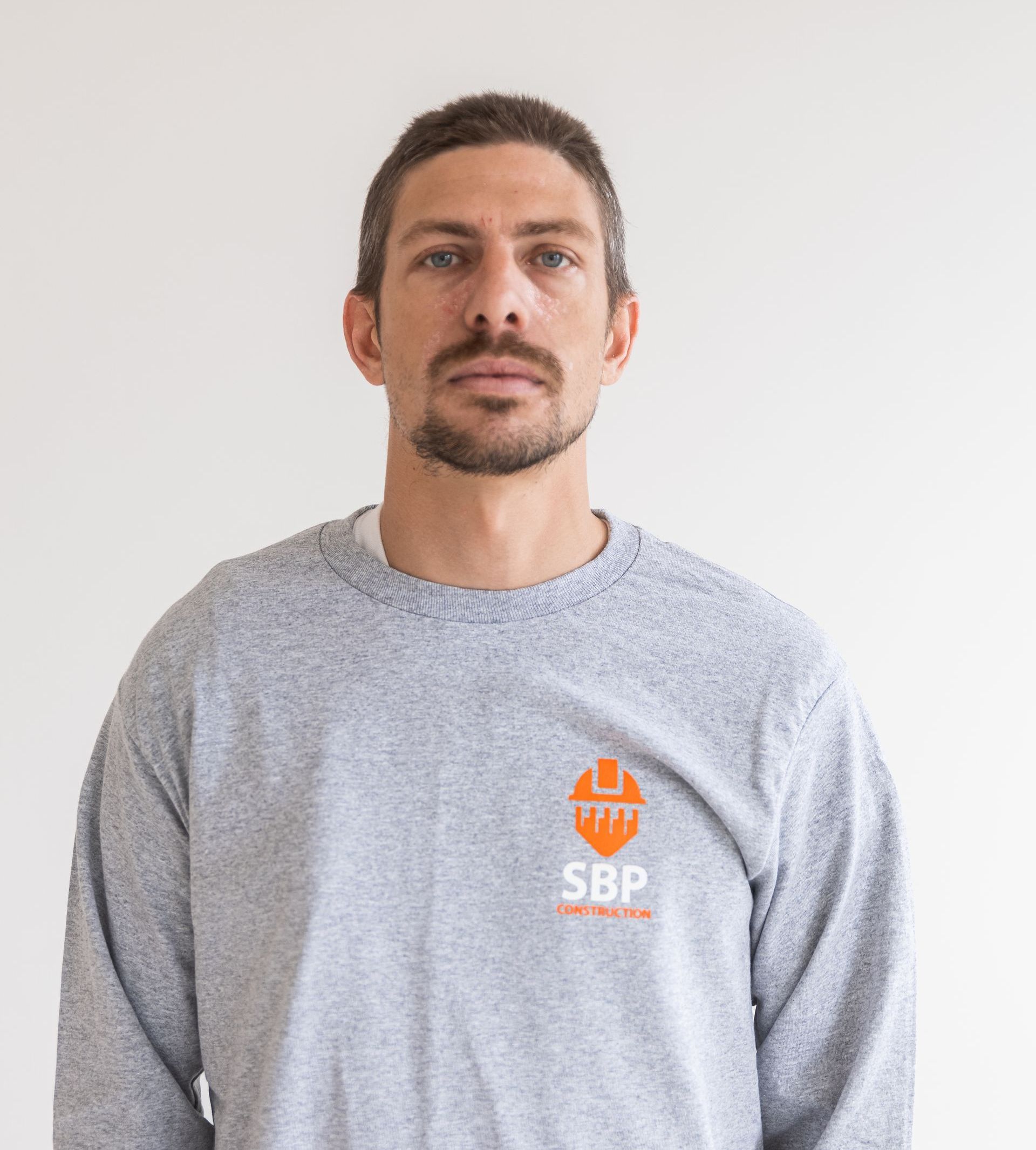 Man in gray sweatshirt with an orange SBP logo. White background.