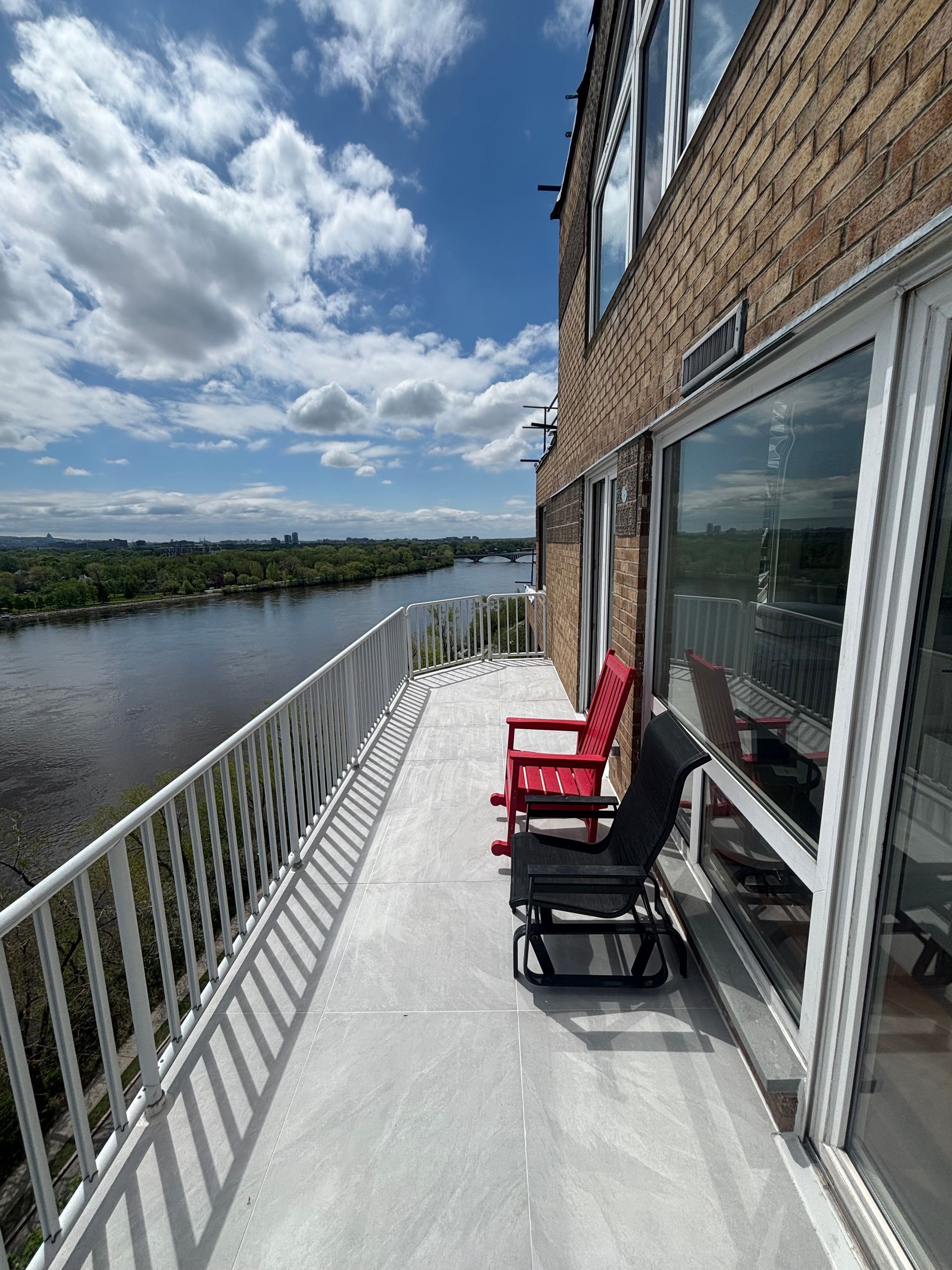 Balcon donnant sur un large fleuve. Des chaises rouges et noires sont disposées sur une surface en béton. Journée ensoleillée avec quelques nuages.