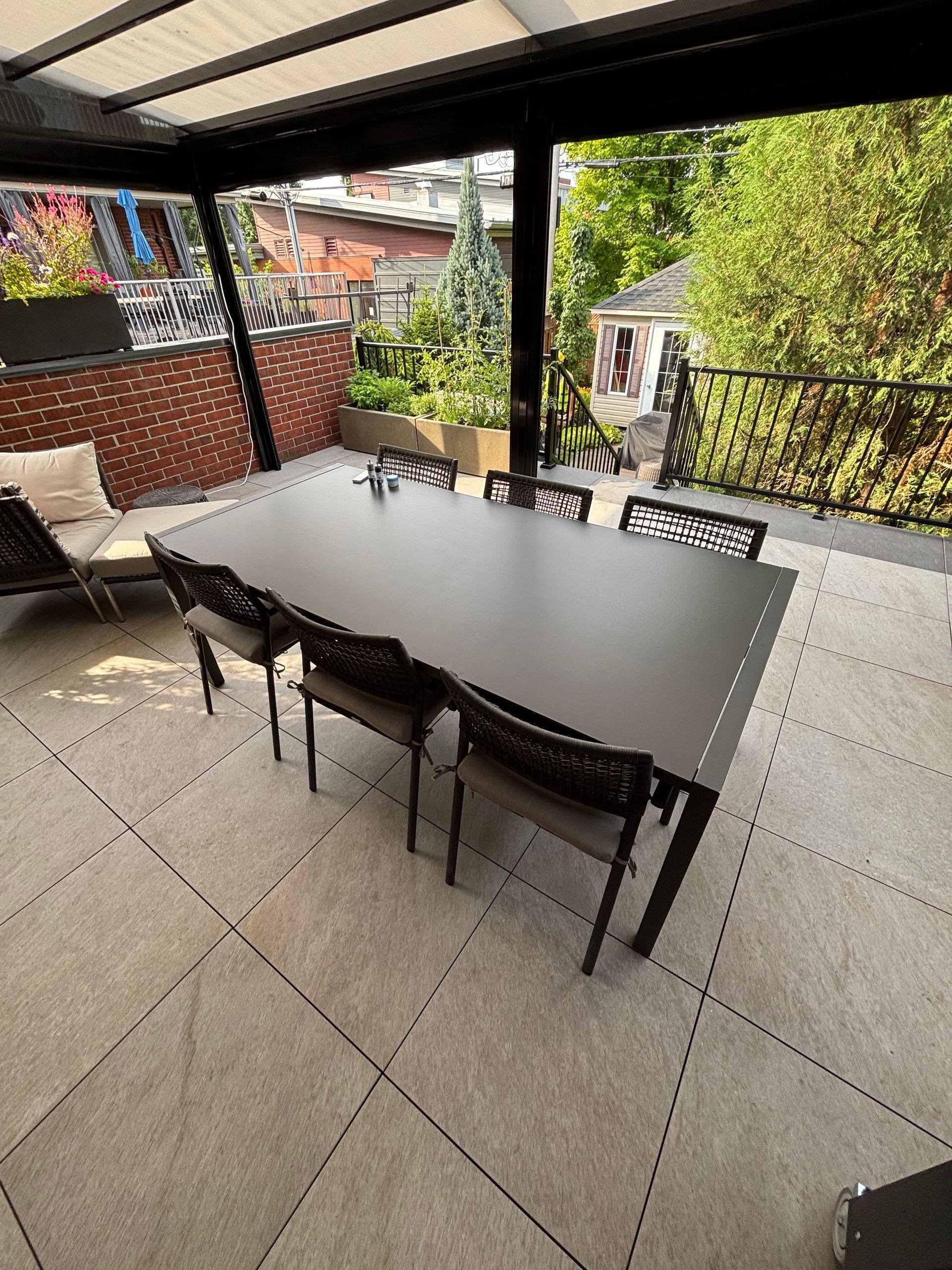 Espace repas extérieur avec une grande table et des chaises noires, sous un auvent sur une terrasse.