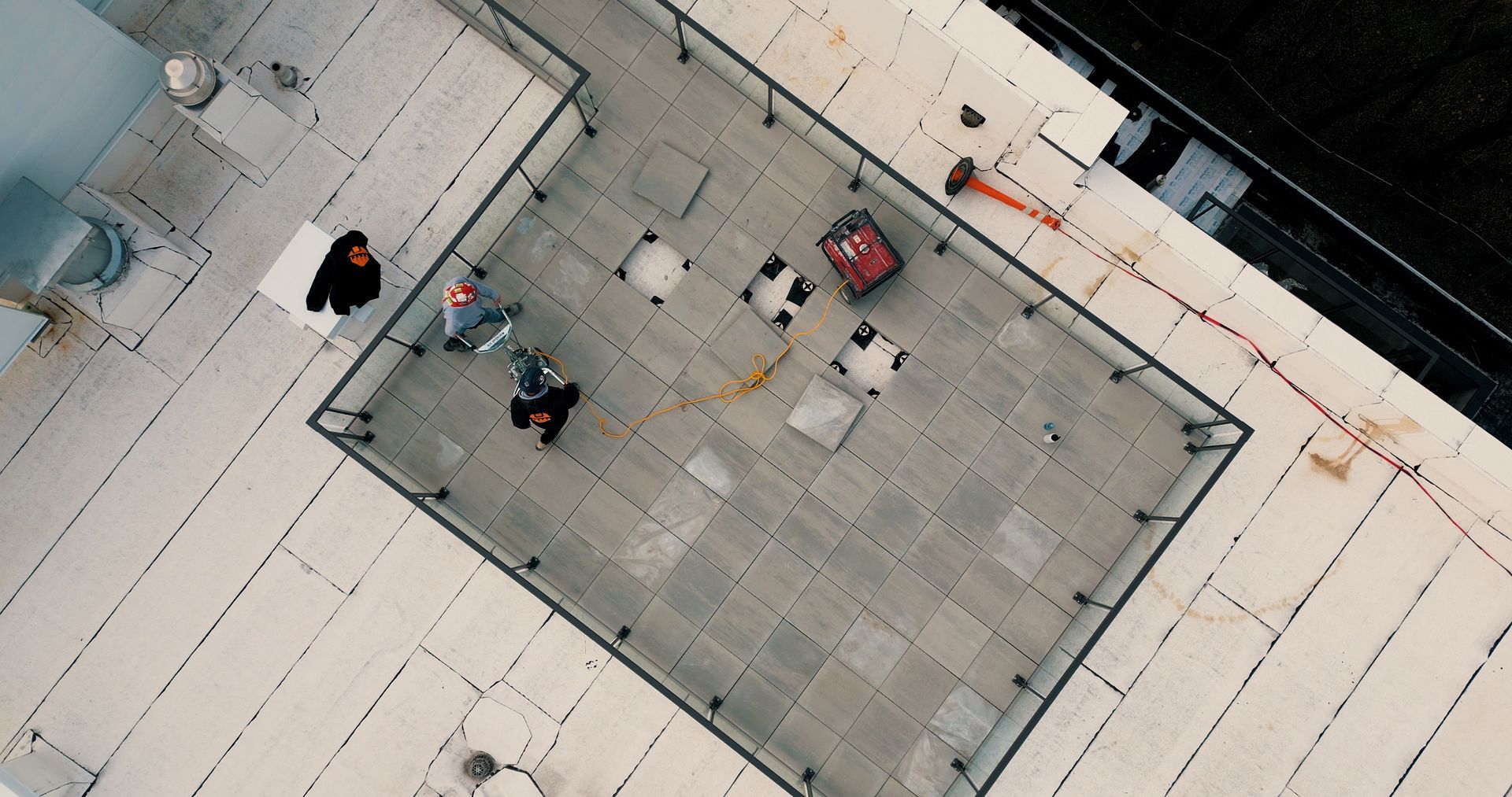 A bird's-eye view of three people working on a rooftop deck with light gray stone tiles and a metal railing.