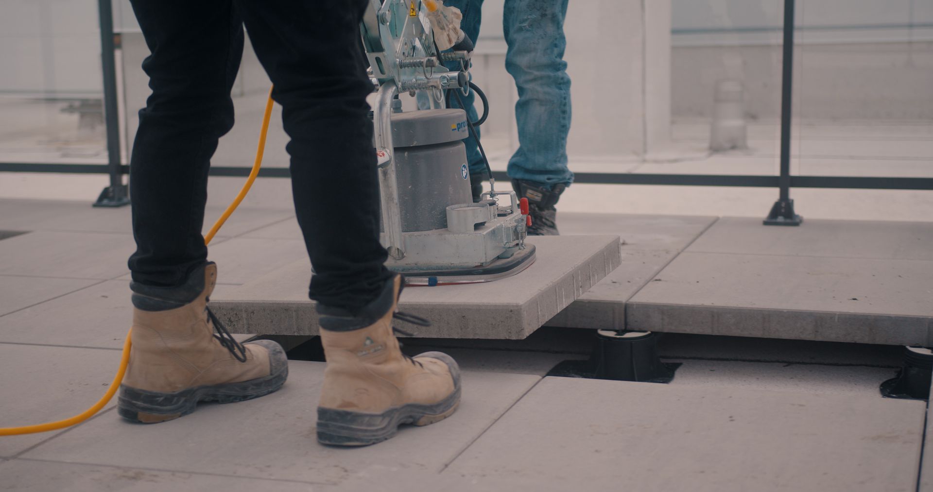 Un ouvrier du bâtiment utilise une meuleuse sur un carreau de pierre à l'extérieur. On aperçoit un câble jaune et des bottes beiges.