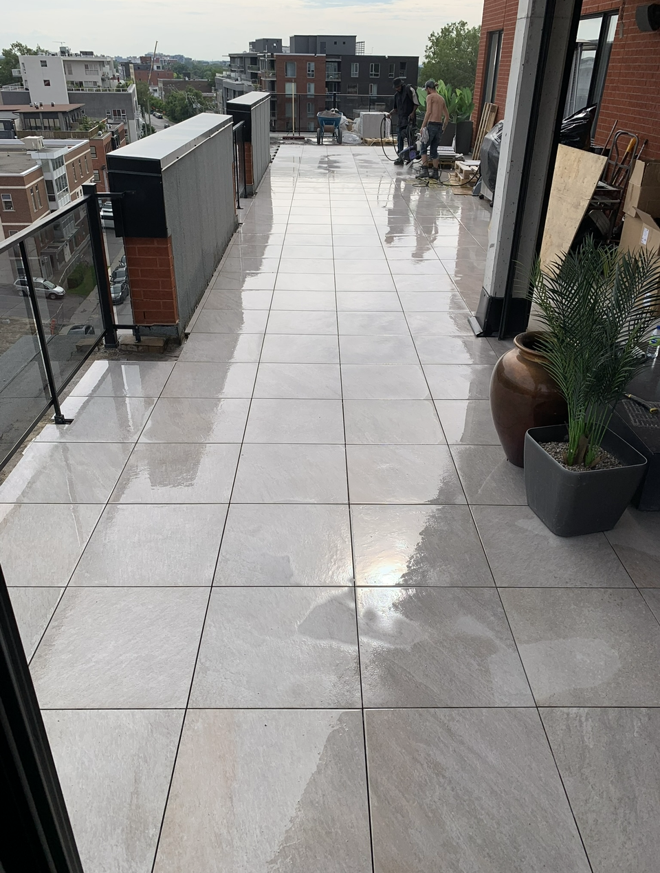 Un balcon au sol carrelé, gris clair et humide, s'étend vers l'extérieur, bordé d'une rambarde et de murets, avec des immeubles de la ville au loin.