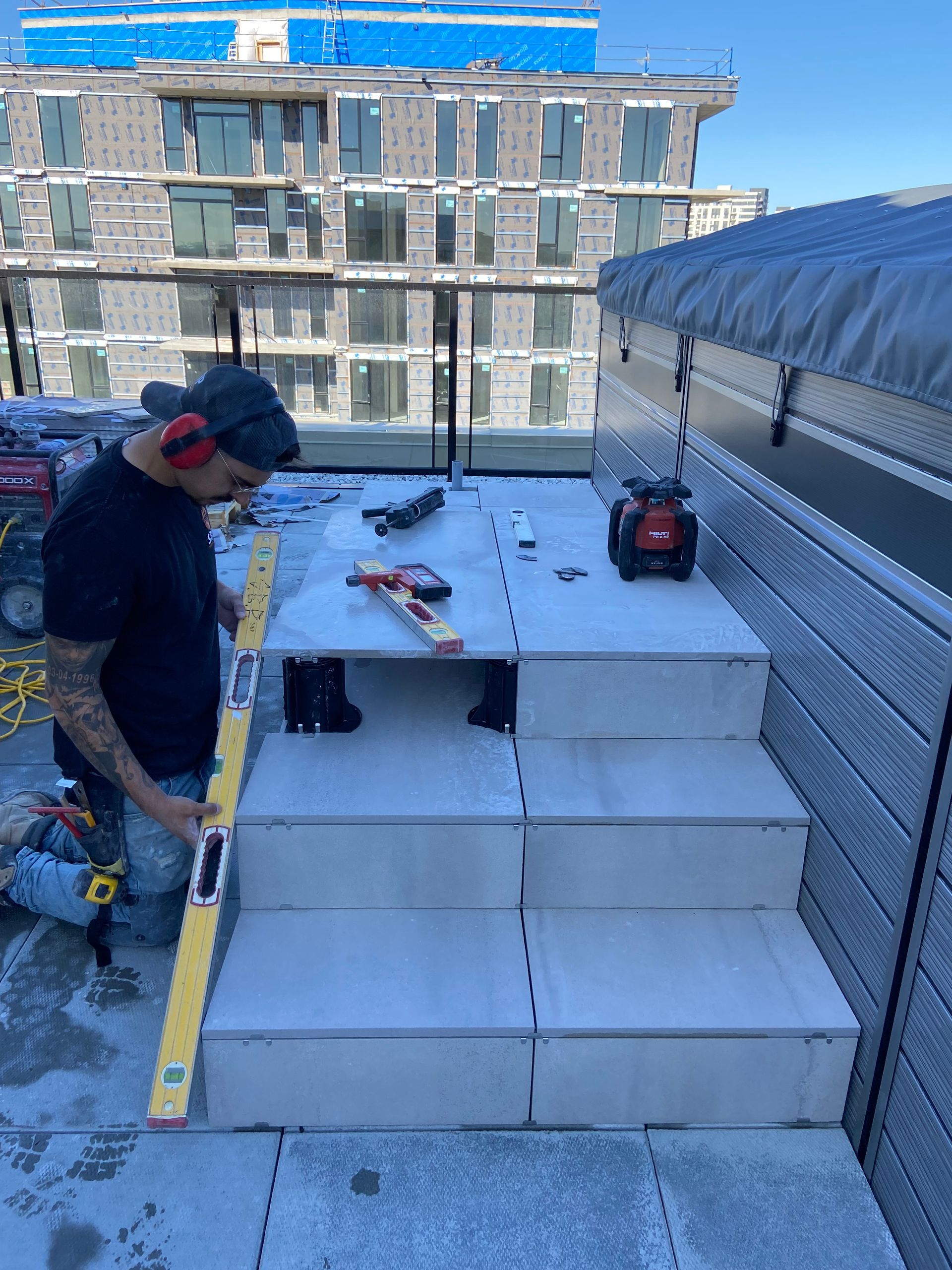 Une personne vêtue d'un t-shirt noir utilise un long niveau jaune pour vérifier l'alignement des marches en mousse blanche sur un balcon extérieur.