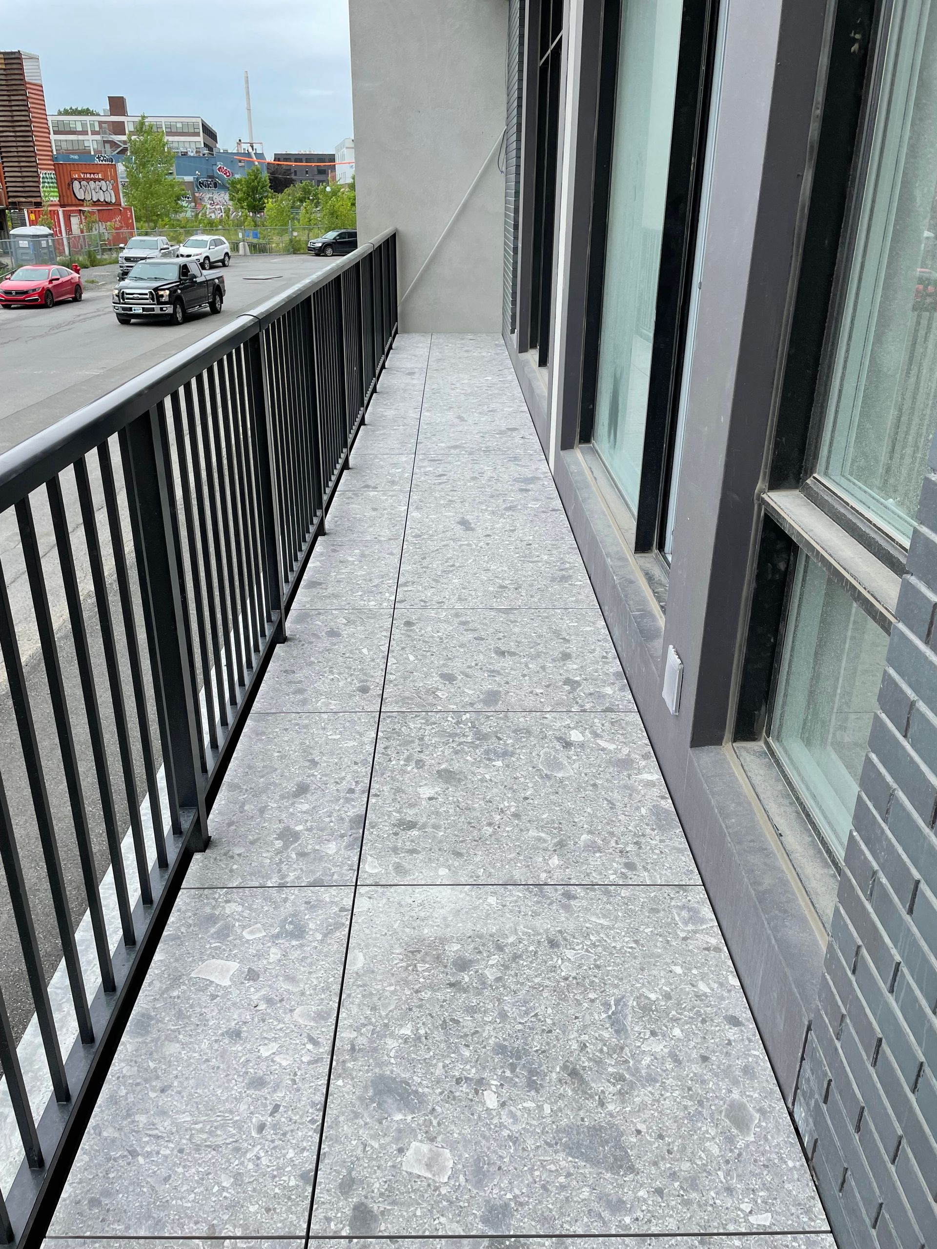 Un balcon long et étroit, au sol carrelé de carreaux gris mouchetés et doté d'une rambarde en métal noir, donnant sur une rue où des voitures sont garées.