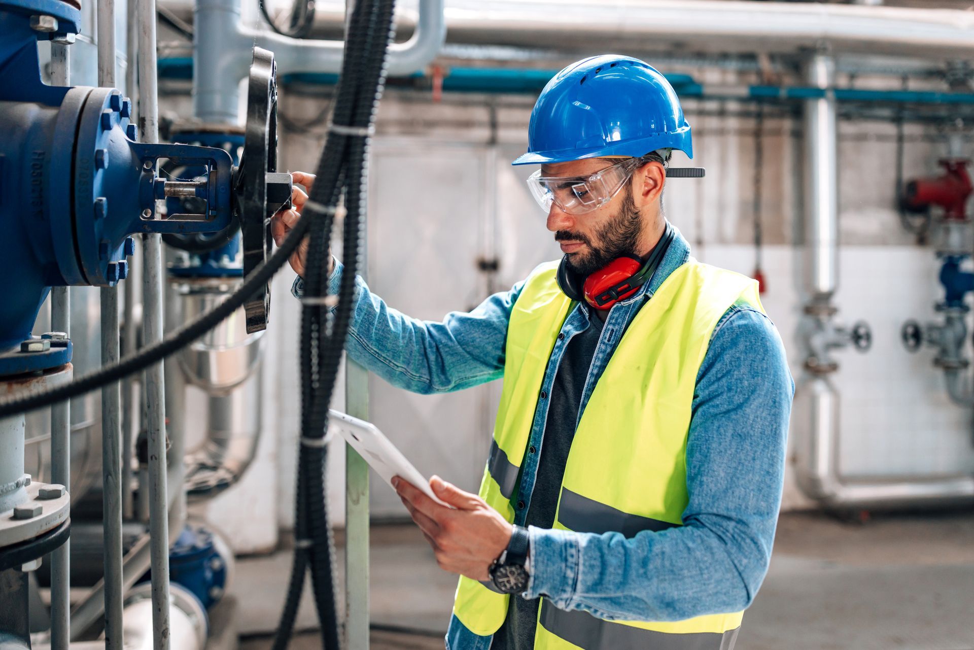 A man in a hard hat and safety vest is looking at a tablet in a factory.