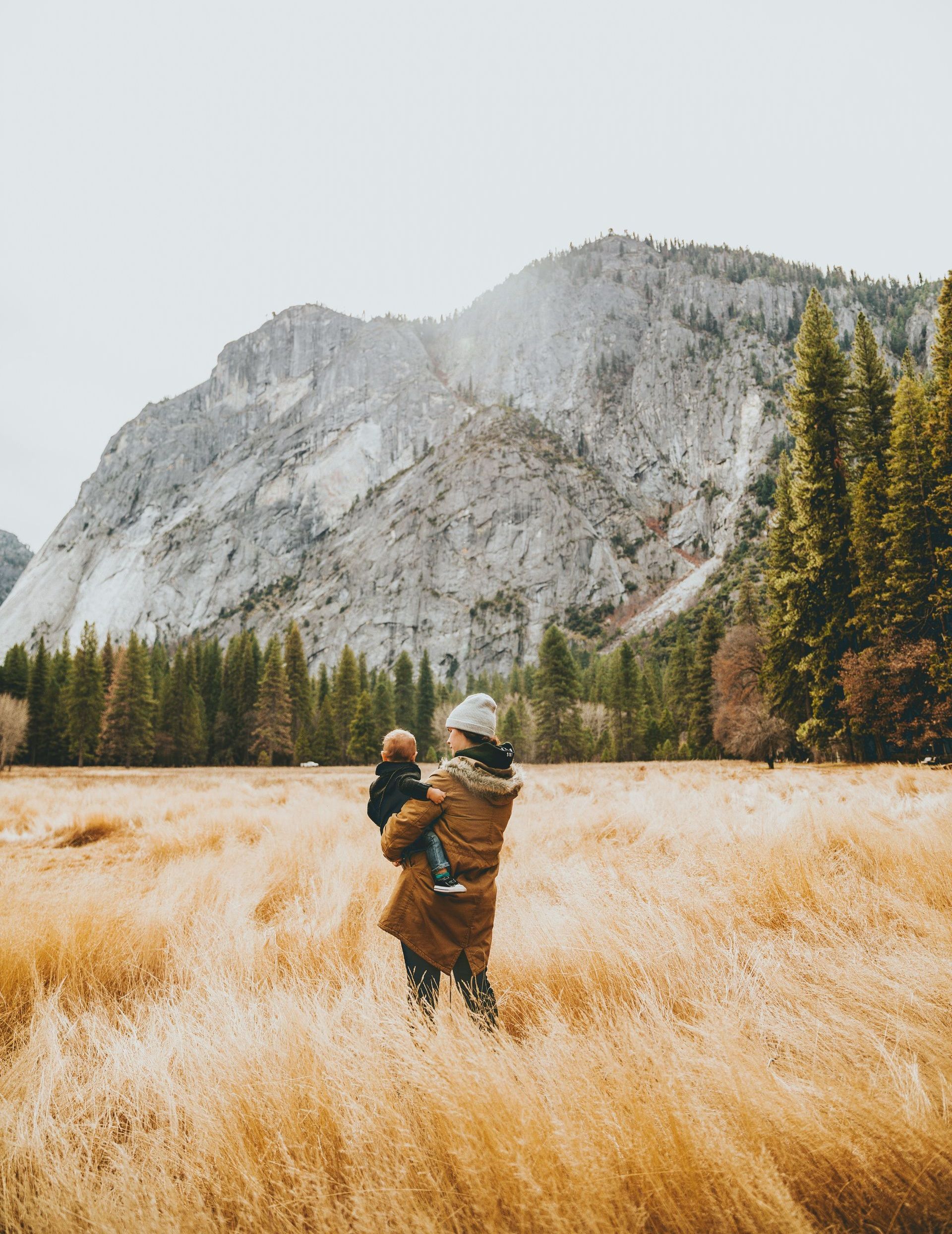 A woman is holding a baby in a field with mountains in the background.
