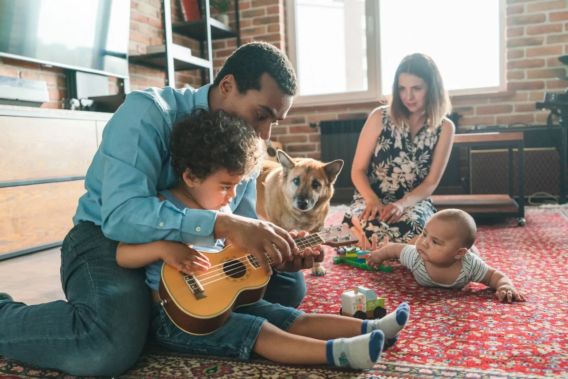 Adult helping child play guitar while a woman and baby sit nearby in a cozy living room with a dog and red rug