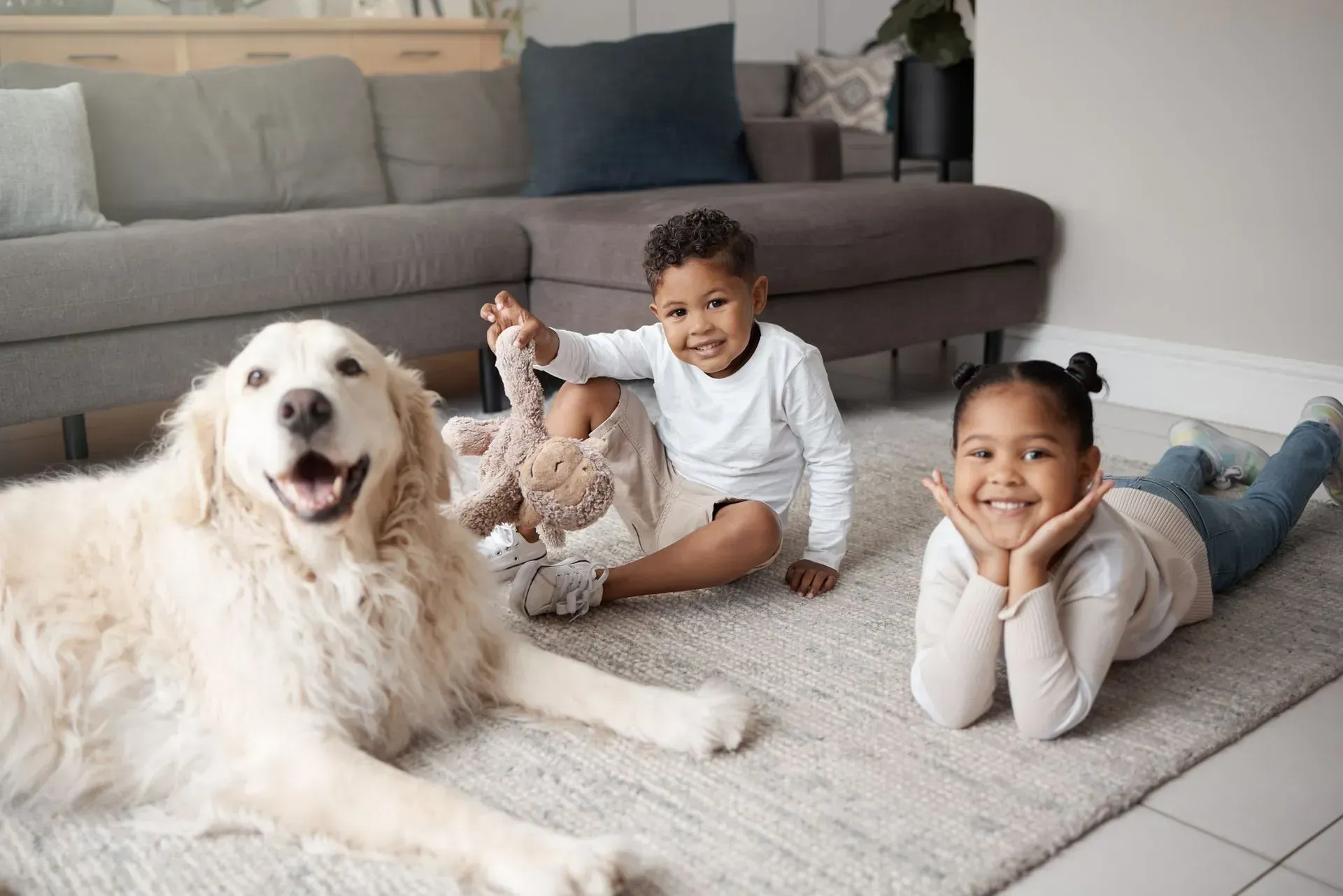 Two children and a fluffy white dog lying on a rug in a bright living room, smiling at the camera