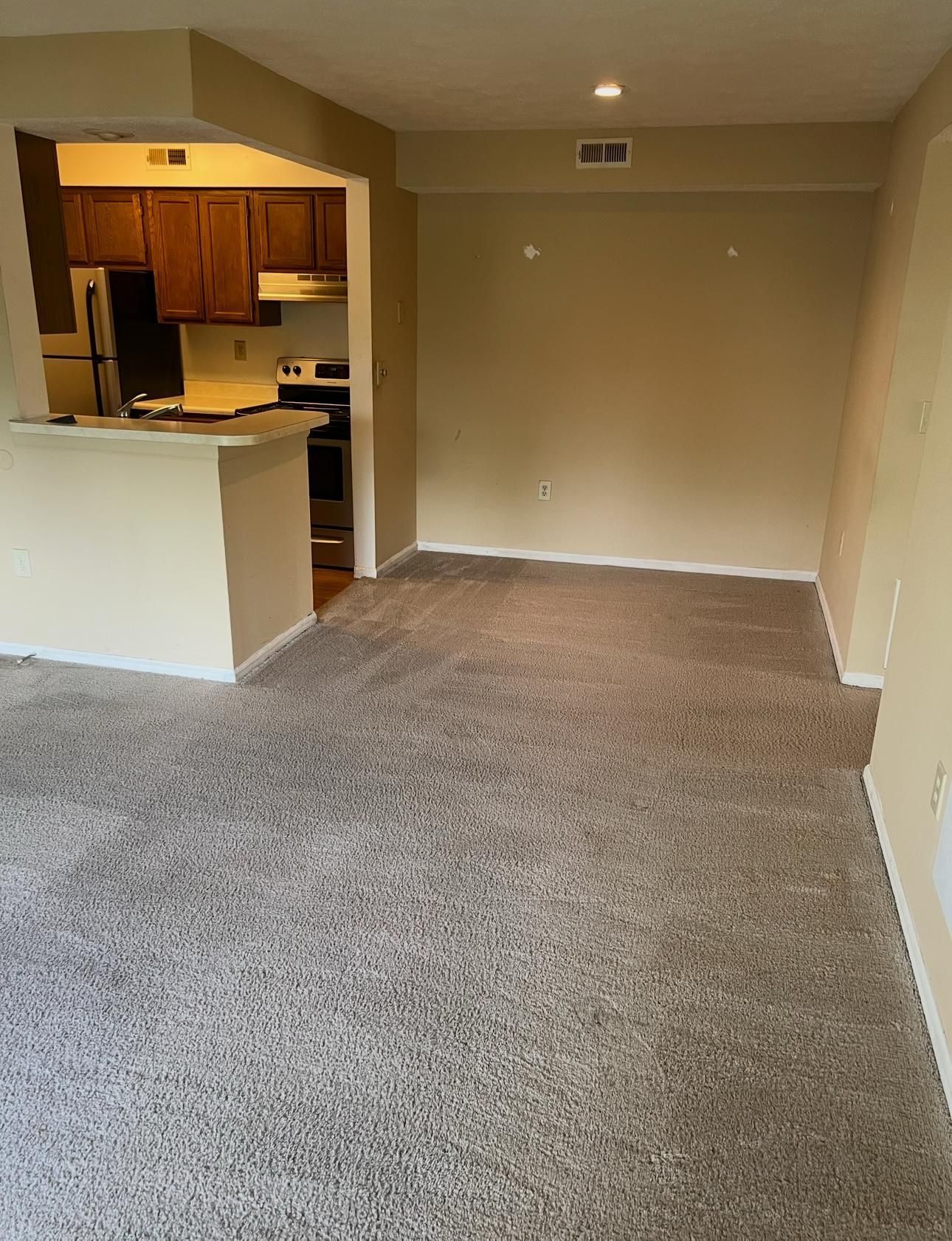 Empty beige living room with carpet, open kitchen on the left, and a doorway at the far end