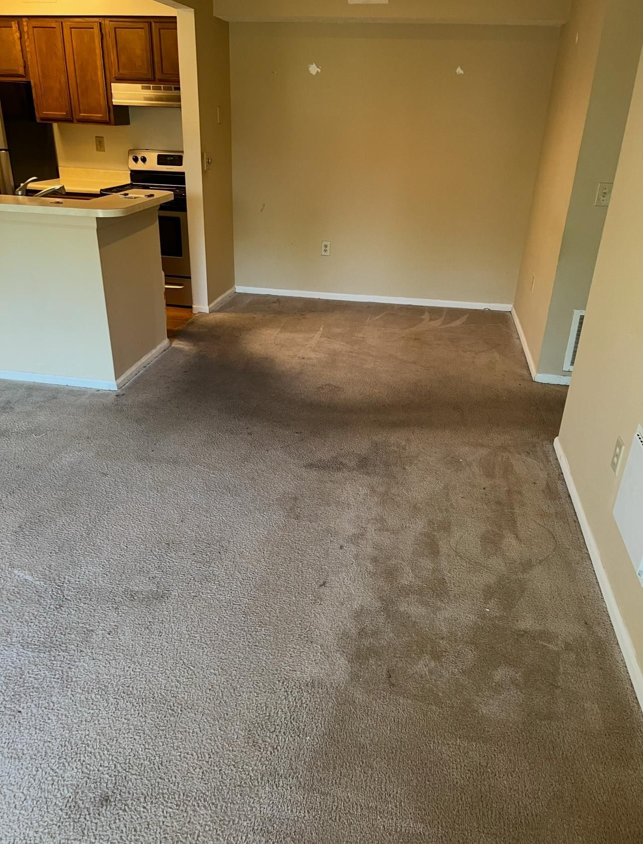 Empty carpeted living room with tan walls and a view into a kitchen area.