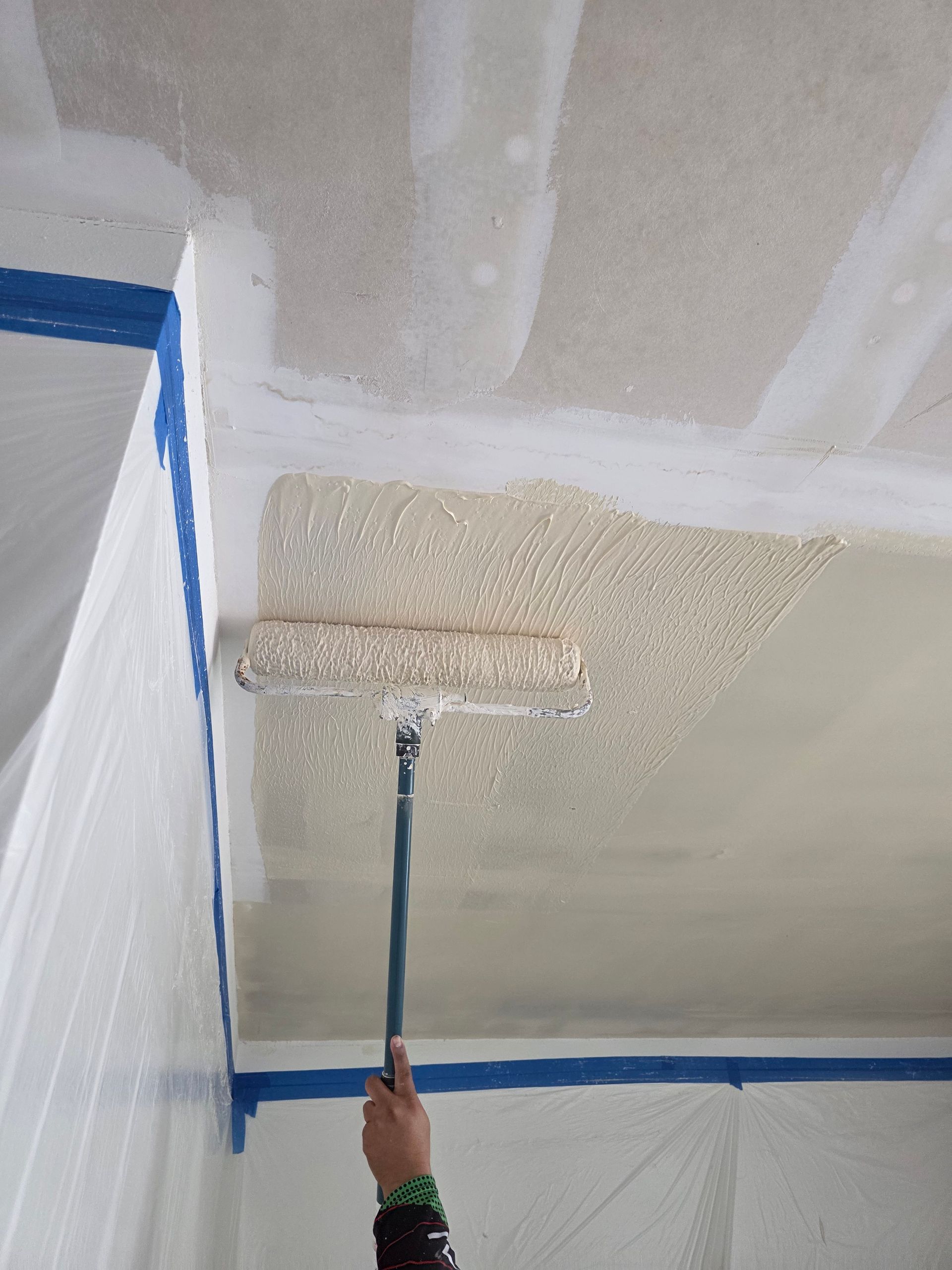 Person painting a ceiling with a roller in a room masked with blue tape and plastic sheeting