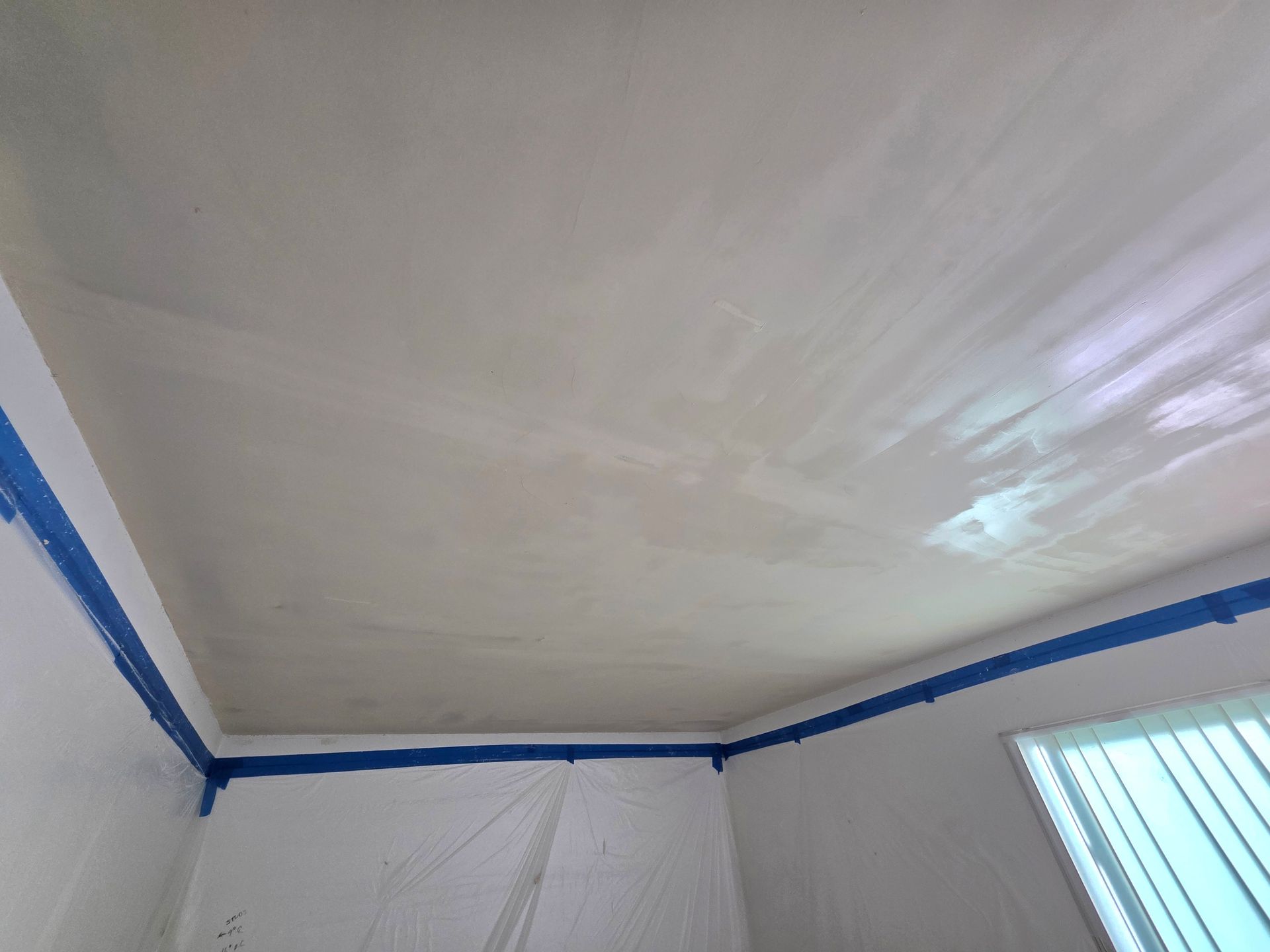 White ceiling and walls with blue painter’s tape along the edges, viewed from a corner near a window.