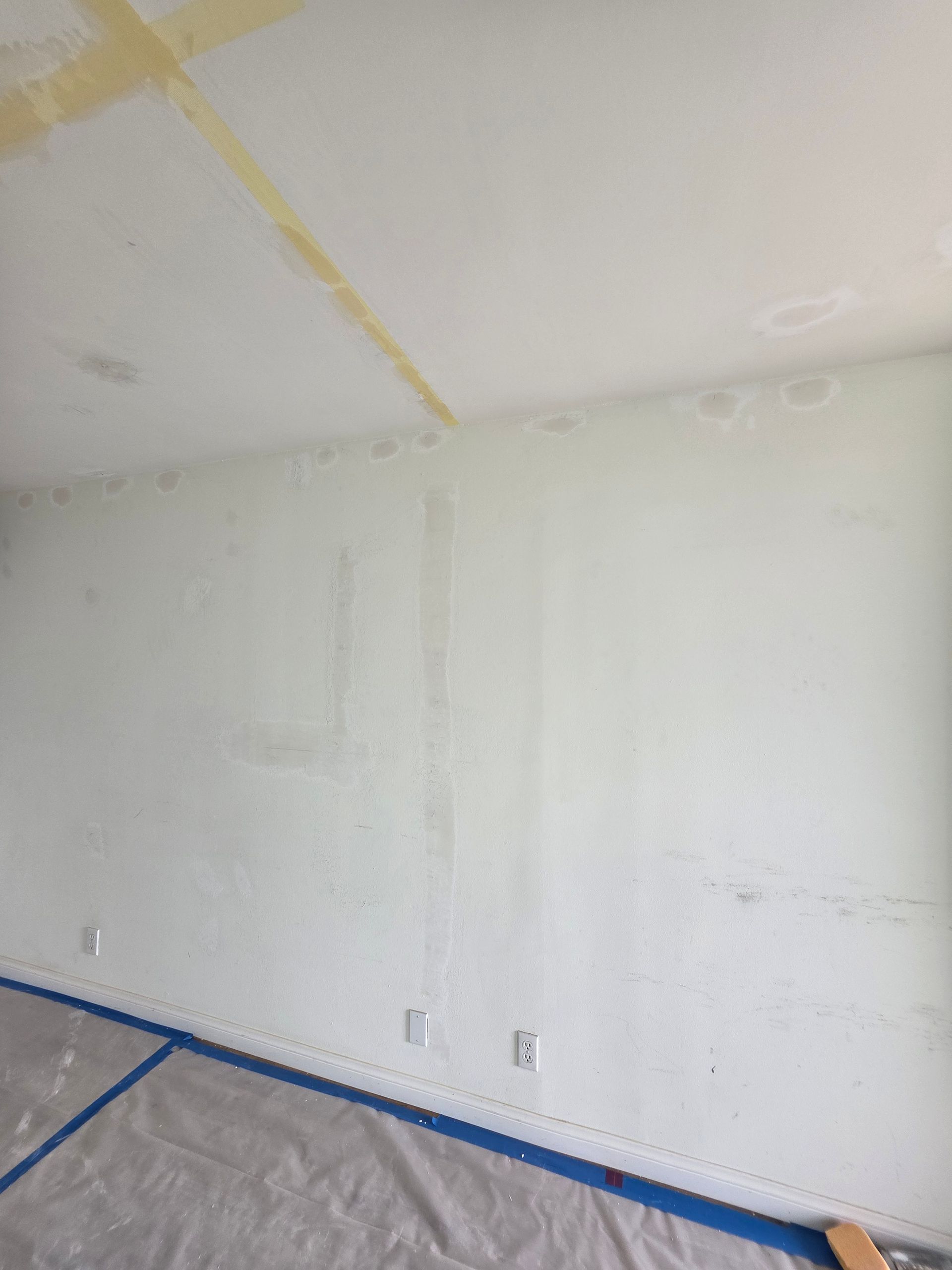Empty white room wall with blue painter’s tape along the floor and plastic sheeting covering the floor.