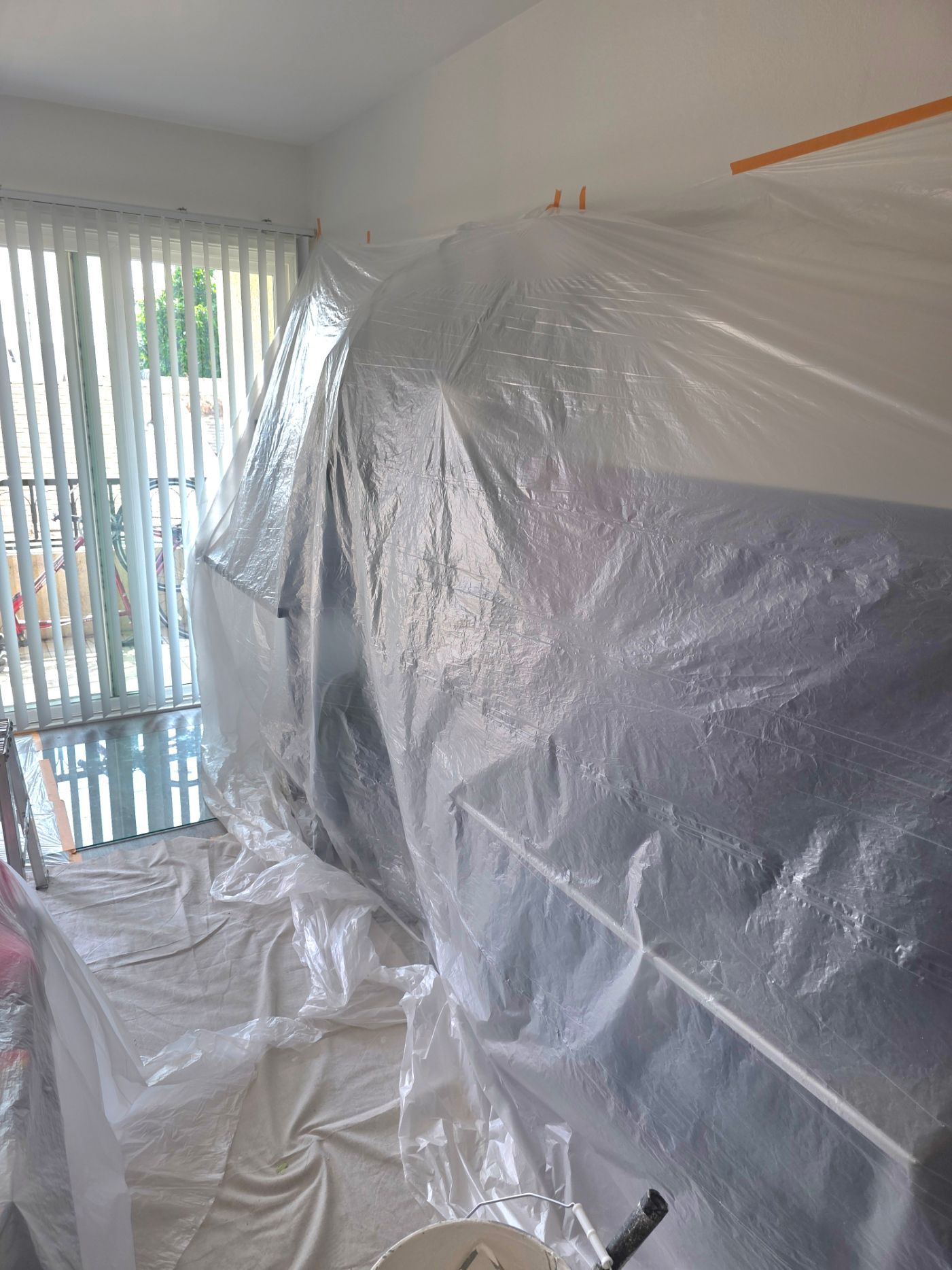 Room covered in plastic sheeting during renovation, with window blinds and a covered sofa.