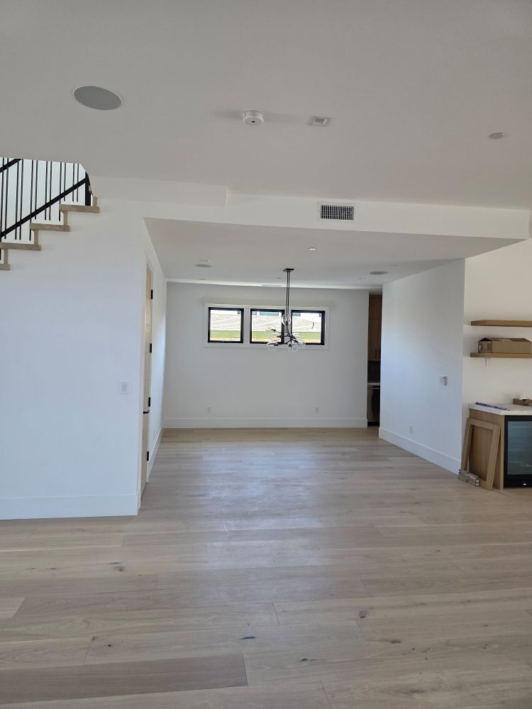 Empty modern living room with light wood floors, white walls, stairs, and three small windows at the far end