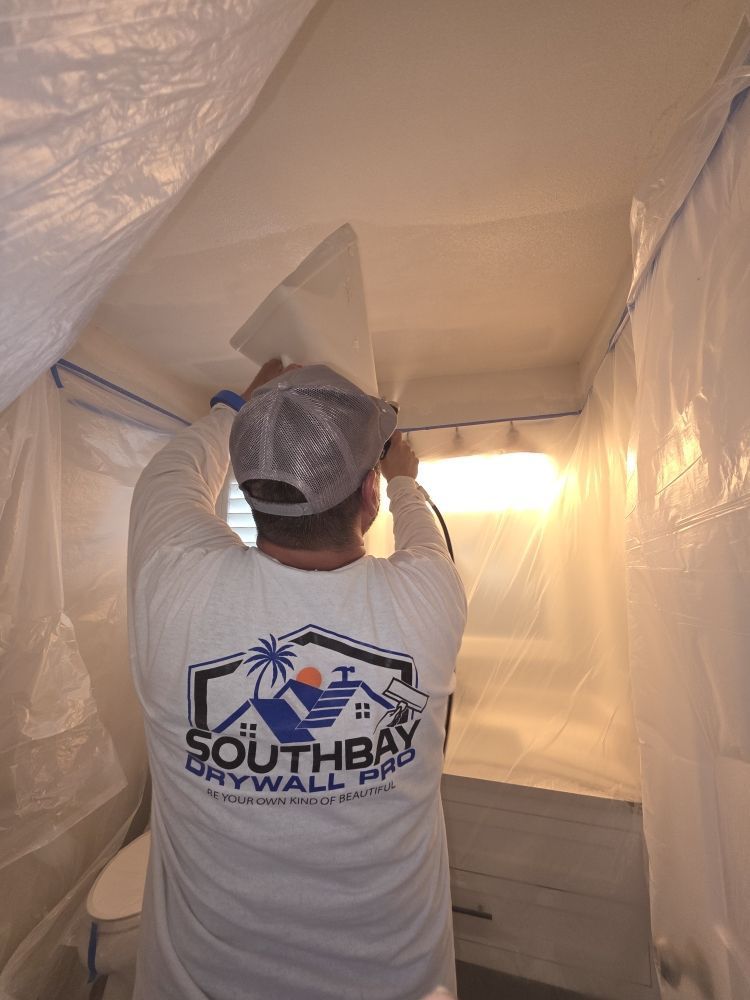 Worker spray-painting a ceiling inside a plastic-covered room, wearing a South Bay Drywall Pro shirt.
