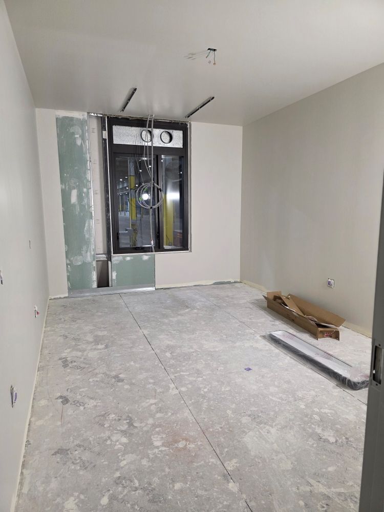 Empty room under renovation with drywall, exposed ceiling fixtures, and construction materials on the floor.