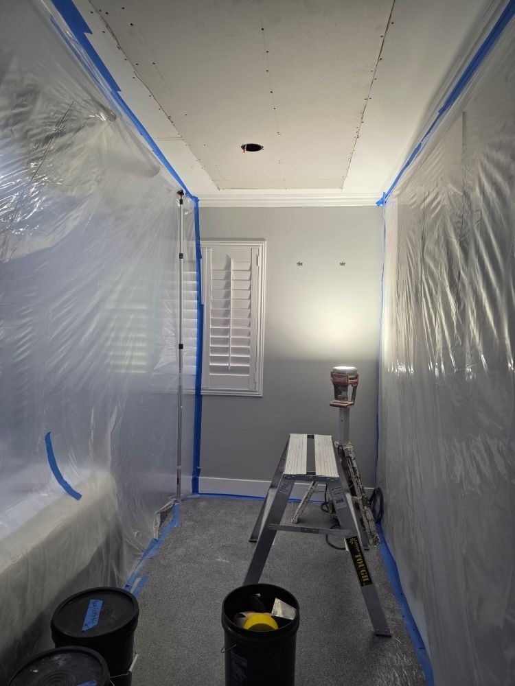 Plastic-covered room under renovation with ladder, paint buckets, and taped window blinds