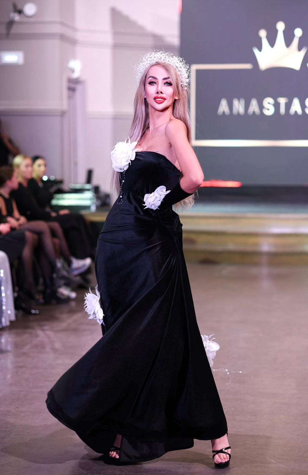 Woman in black dress and headpiece, modeling on runway. White accents, smiling.