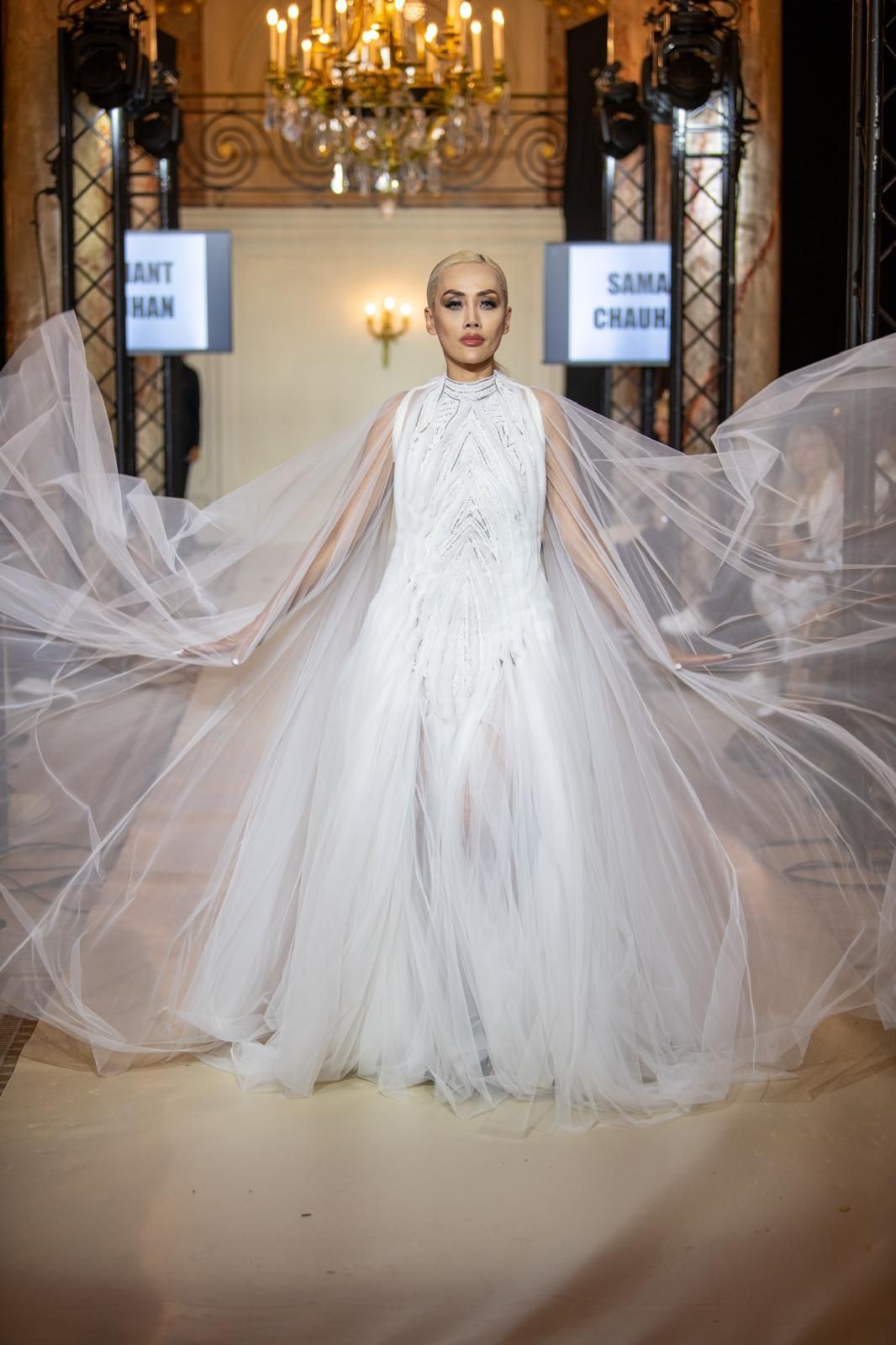 Woman in a flowing white gown on a runway; translucent cape, elaborate bodice, setting with lights, signs.