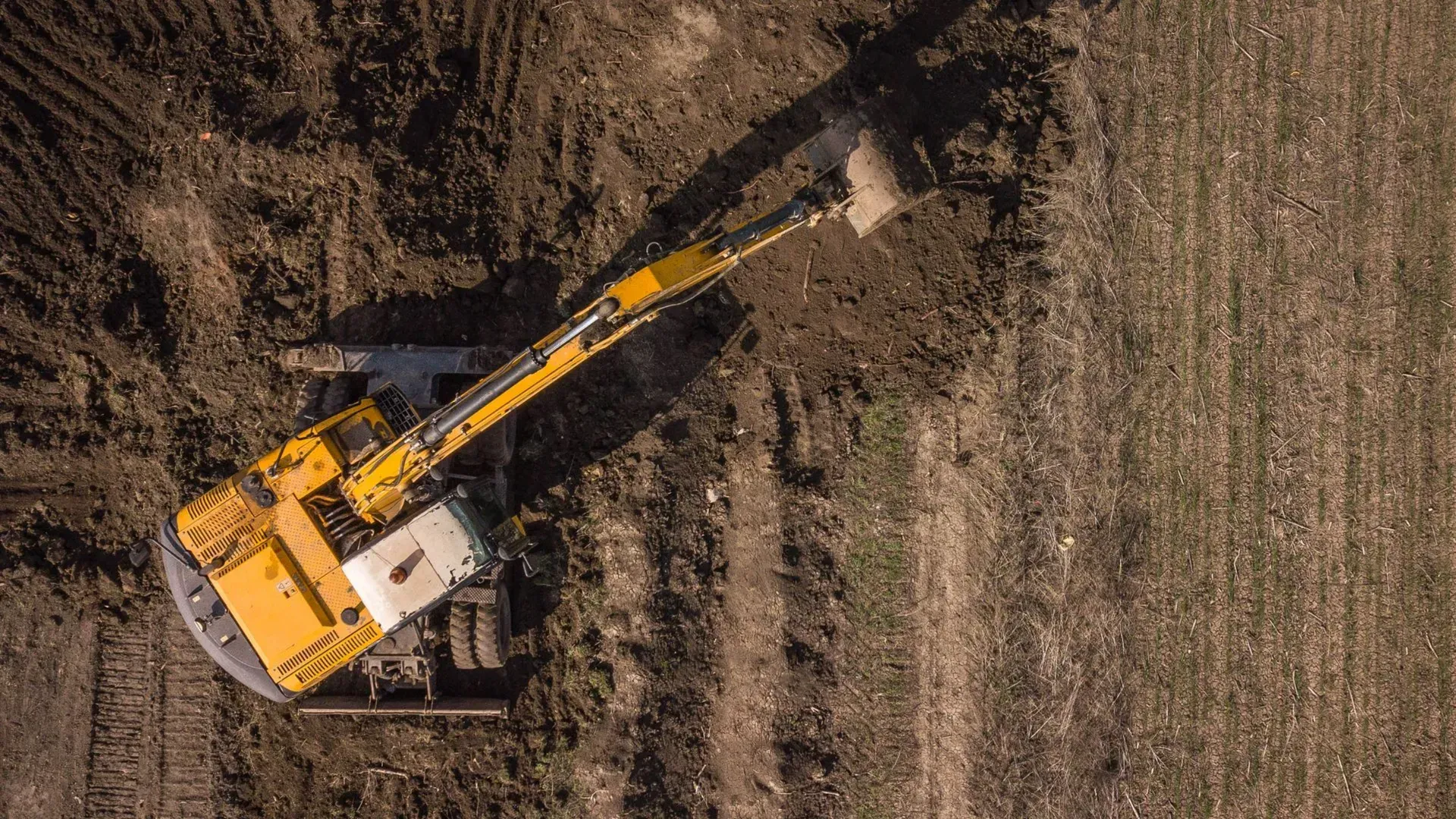 Une excavatrice jaune dans un champ de terre, vue du ciel, avec son bras étendu au-dessus d'une bande dégagée.