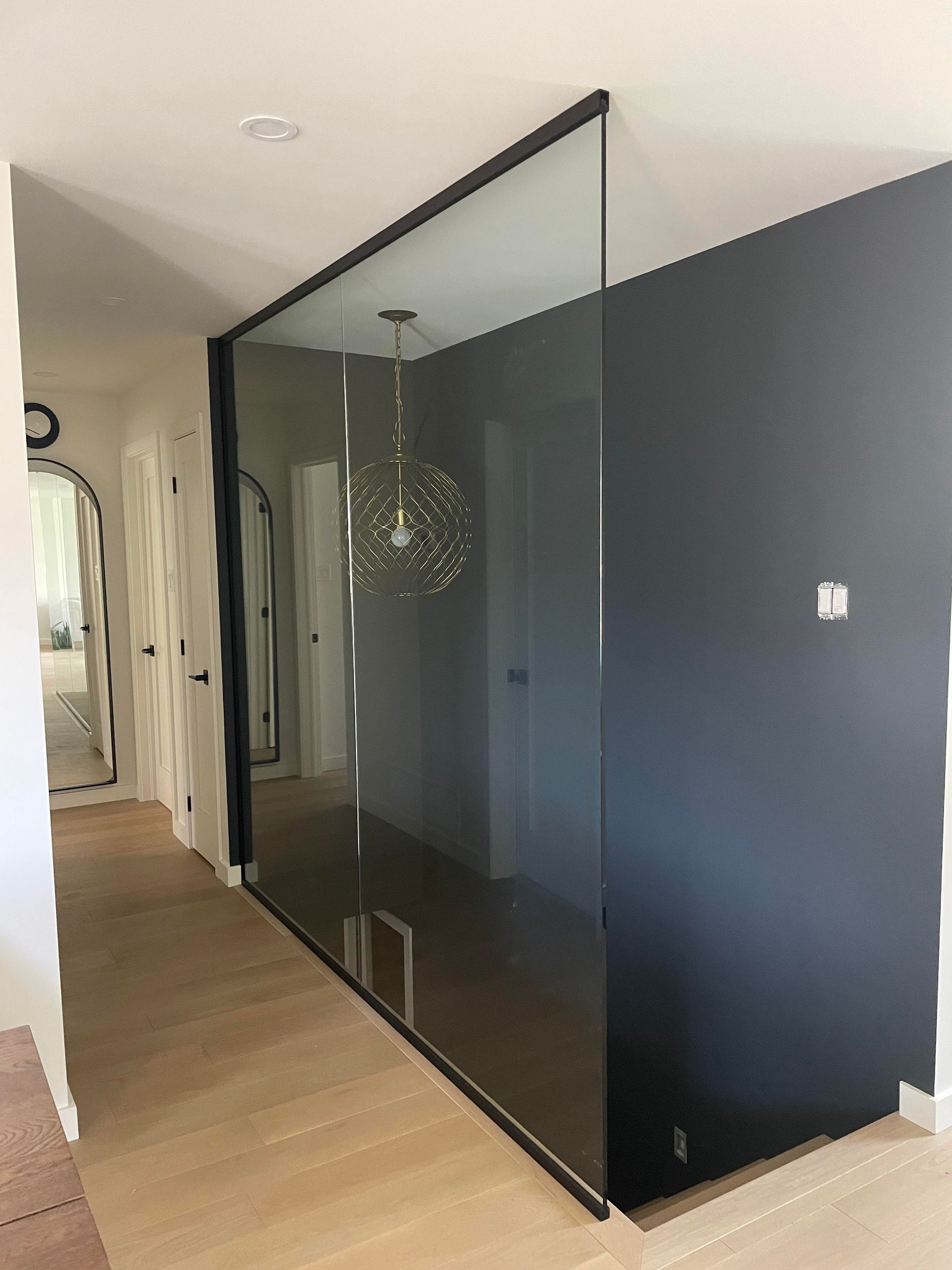 Un couloir avec une paroi vitrée divisant l'espace, un mur gris foncé à droite, un parquet clair et un lustre au loin.