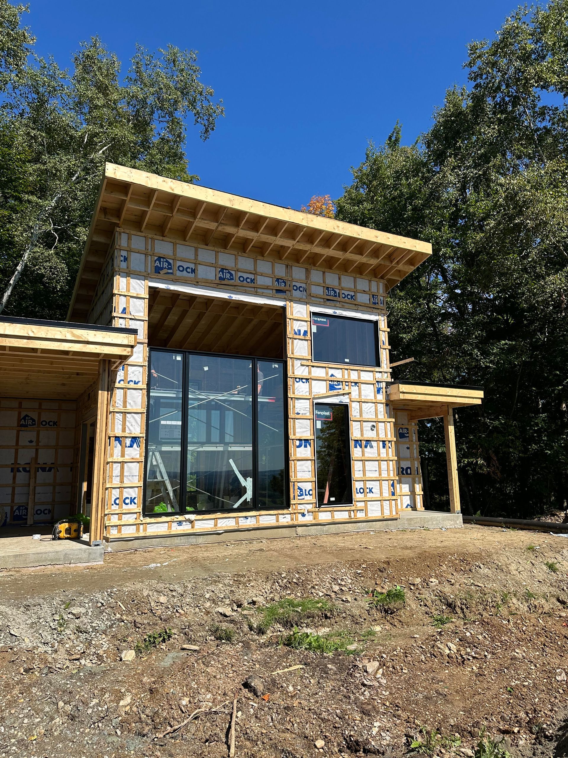 Construction d'une maison moderne, grandes fenêtres, ossature bois, ciel bleu, entourée d'arbres.