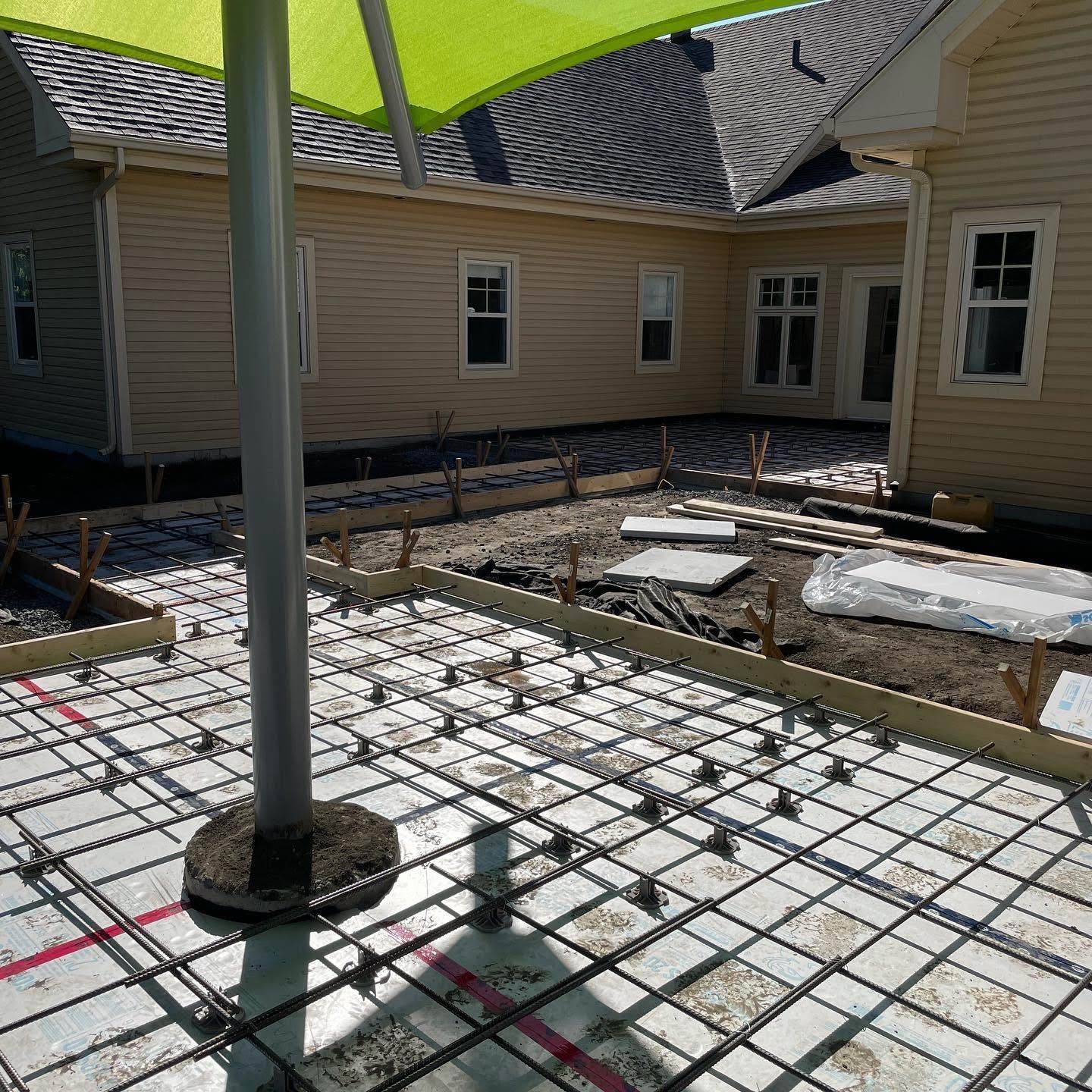 Terrasse en béton en construction ; treillis d'armature, coffrages en bois, mât de parasol, maison beige en arrière-plan.