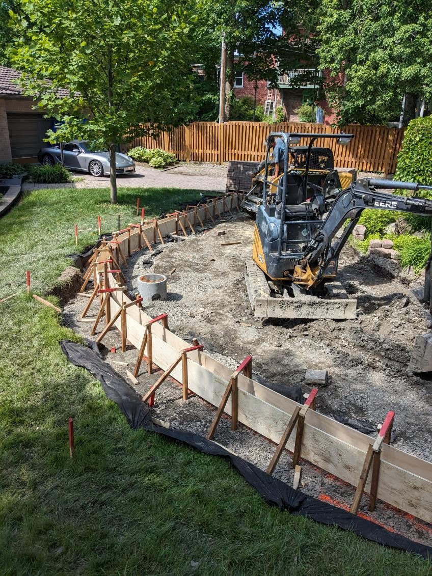Construction d'une bordure en béton incurvée dans une cour résidentielle, avec une petite excavatrice présente.