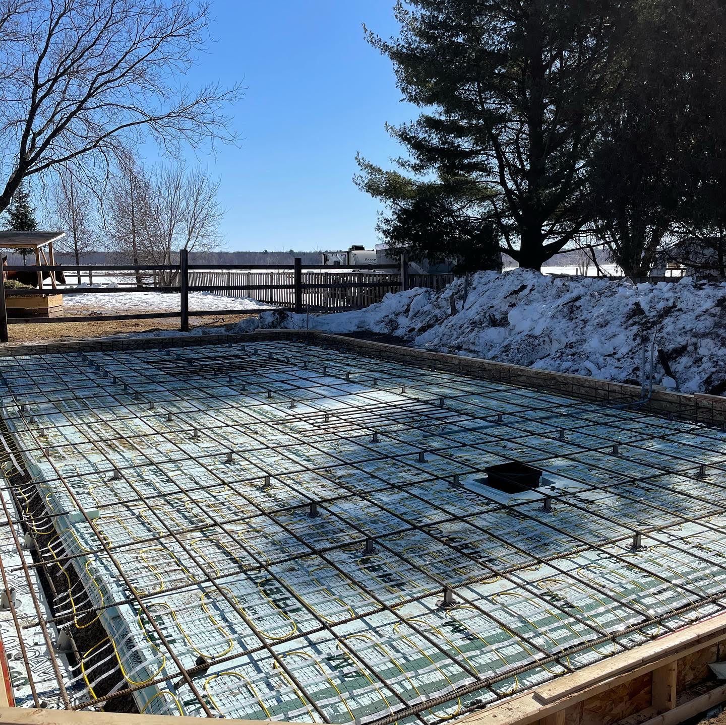 Chantier de construction avec treillis d'armature apparent, isolation en mousse et ossature bois, sol enneigé en arrière-plan sous un ciel bleu.