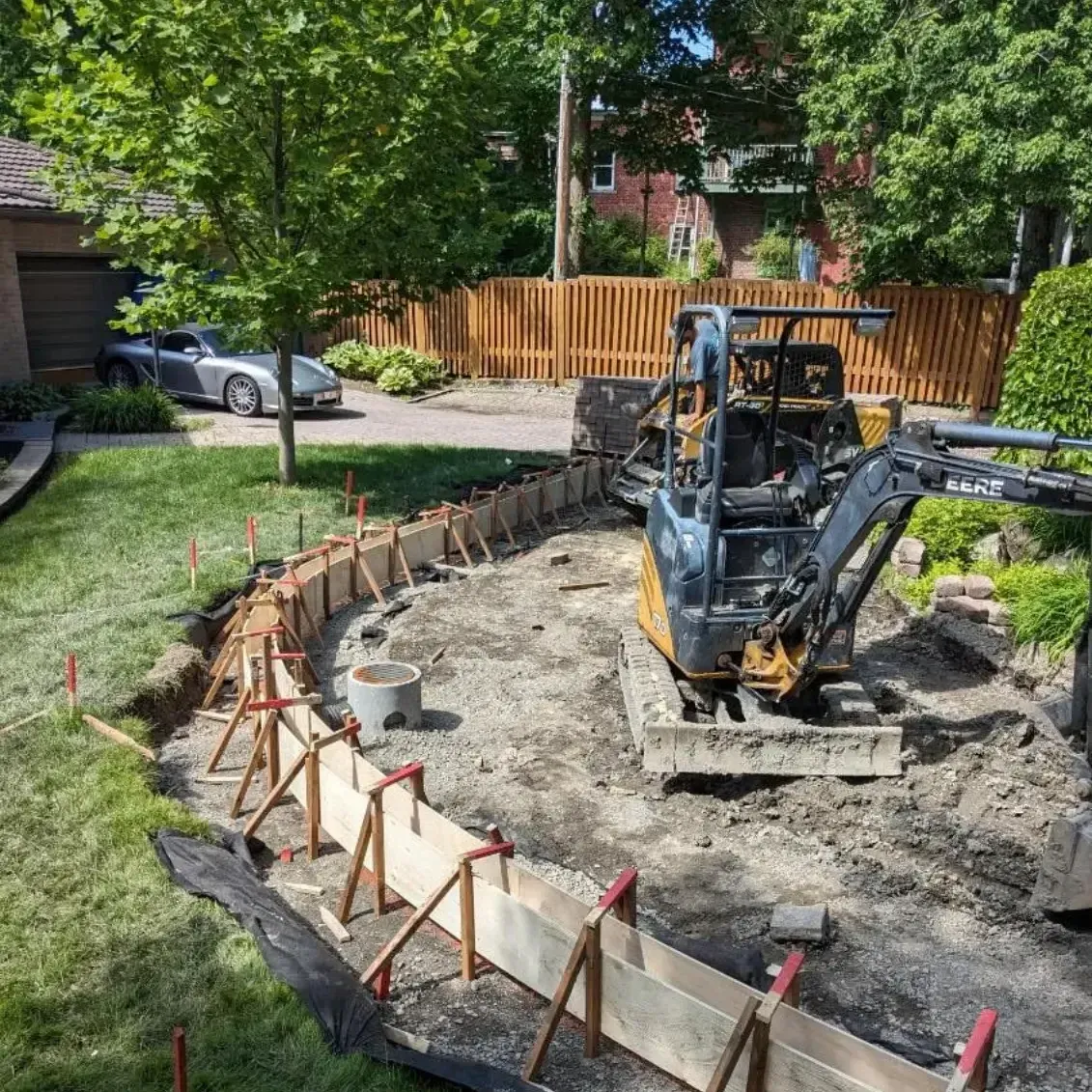 Une pelleteuse creuse une tranchée de fondation incurvée dans un jardin, près d'une clôture en bois et d'arbres.