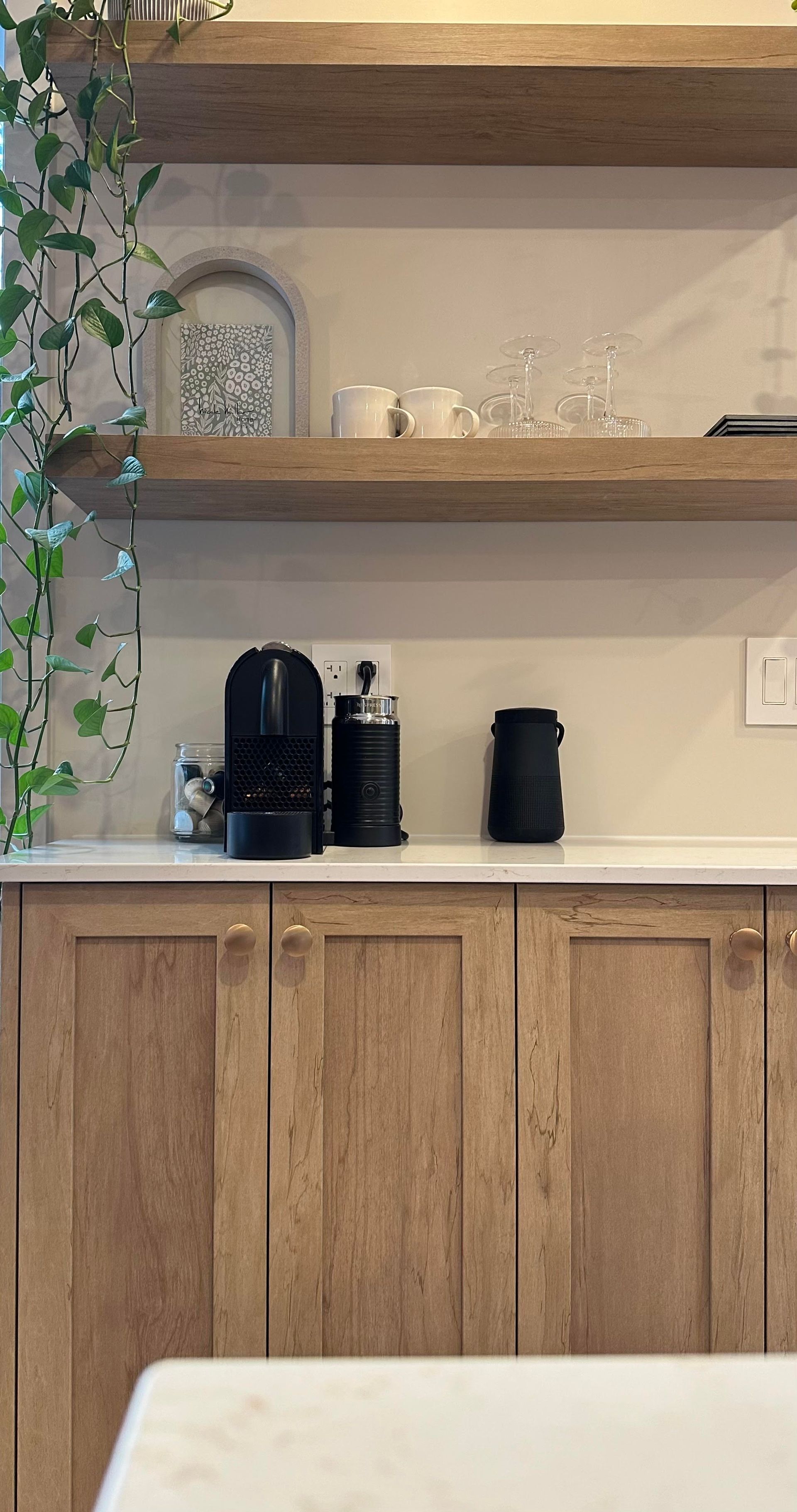 Cuisine avec placards en bois, étagères flottantes et cafetière. Du lierre grimpe au mur.