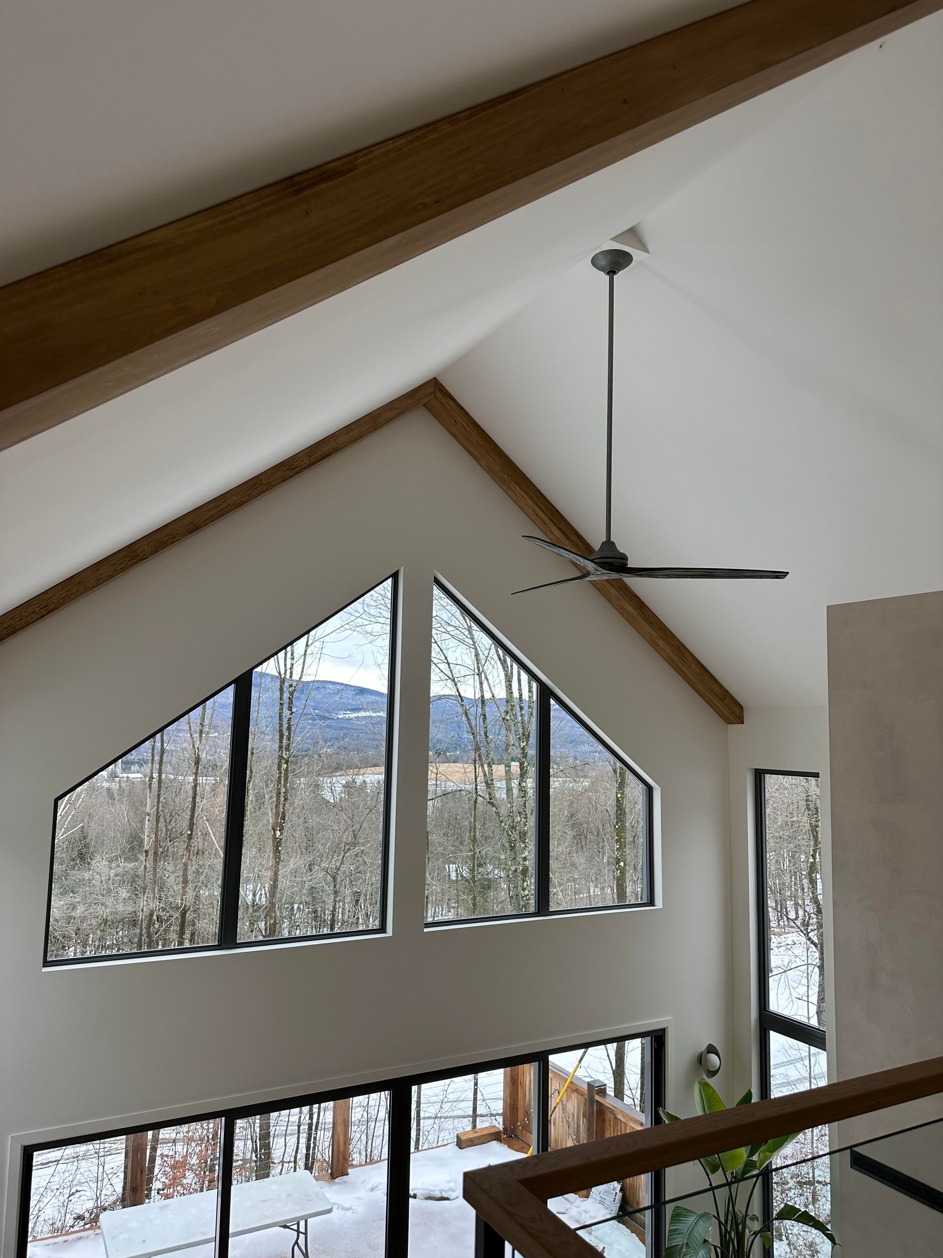 Pièce à haut plafond avec fenêtres triangulaires donnant sur des arbres enneigés. Un ventilateur de plafond est suspendu au toit en pente.