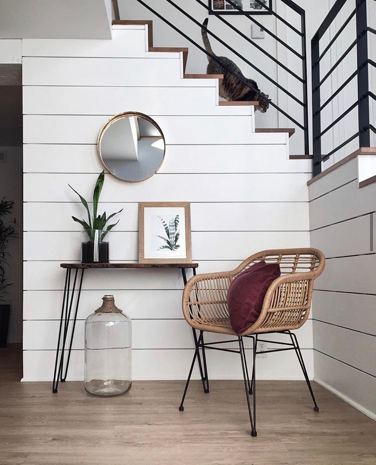 Intérieur avec mur lambrissé blanc, escalier avec chat, table, miroir, plante, chaise et grande carafe en verre.