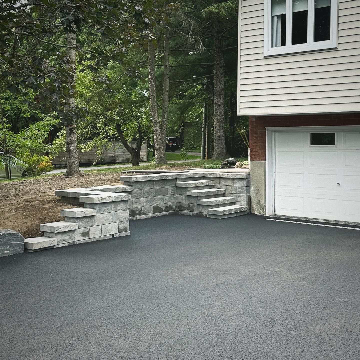 Allée asphaltée avec marches et muret en pierre, jouxtant une maison avec une porte de garage blanche.