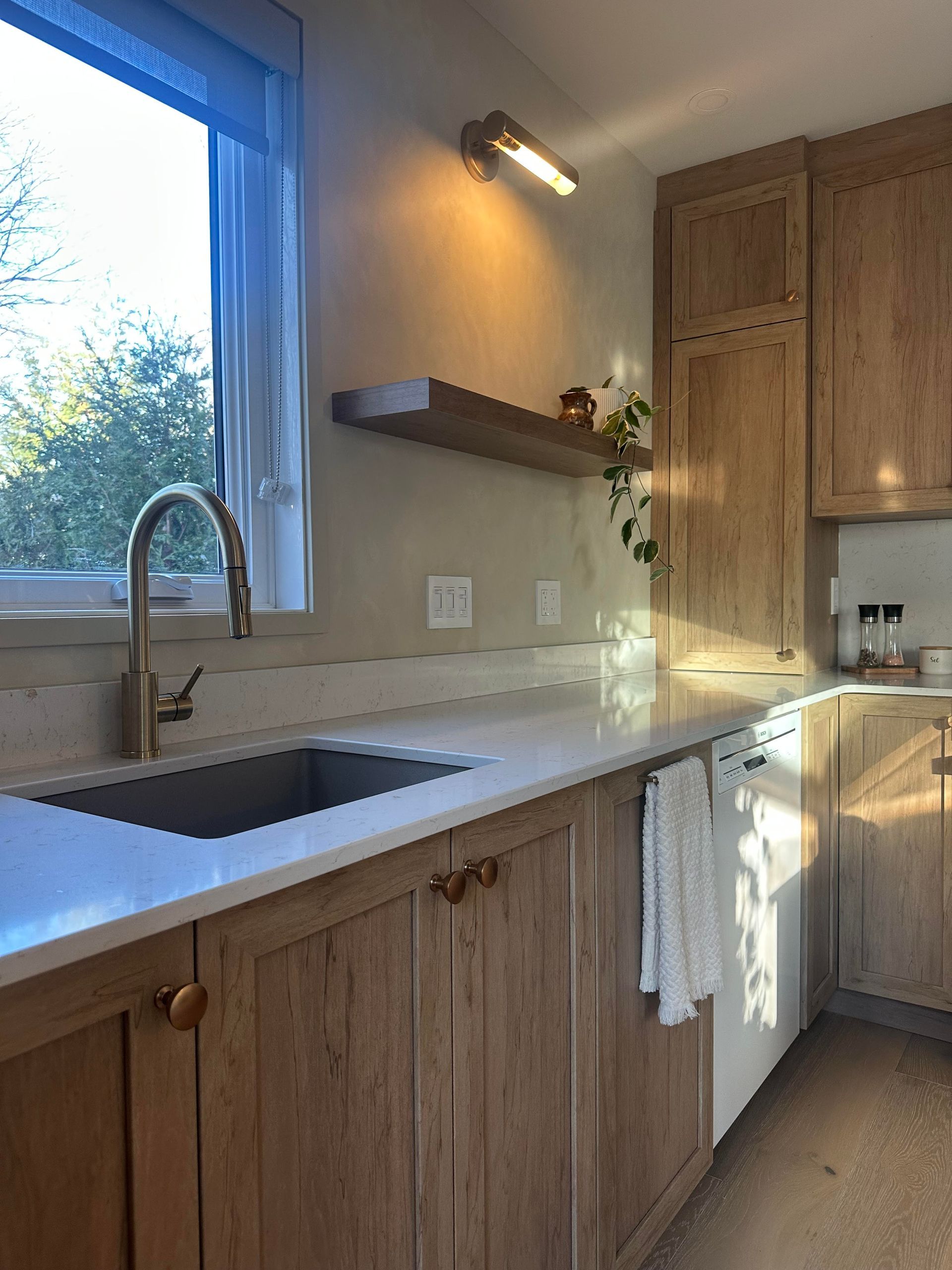 Cuisine avec placards en bois clair, plan de travail blanc, évier en inox et fenêtre.