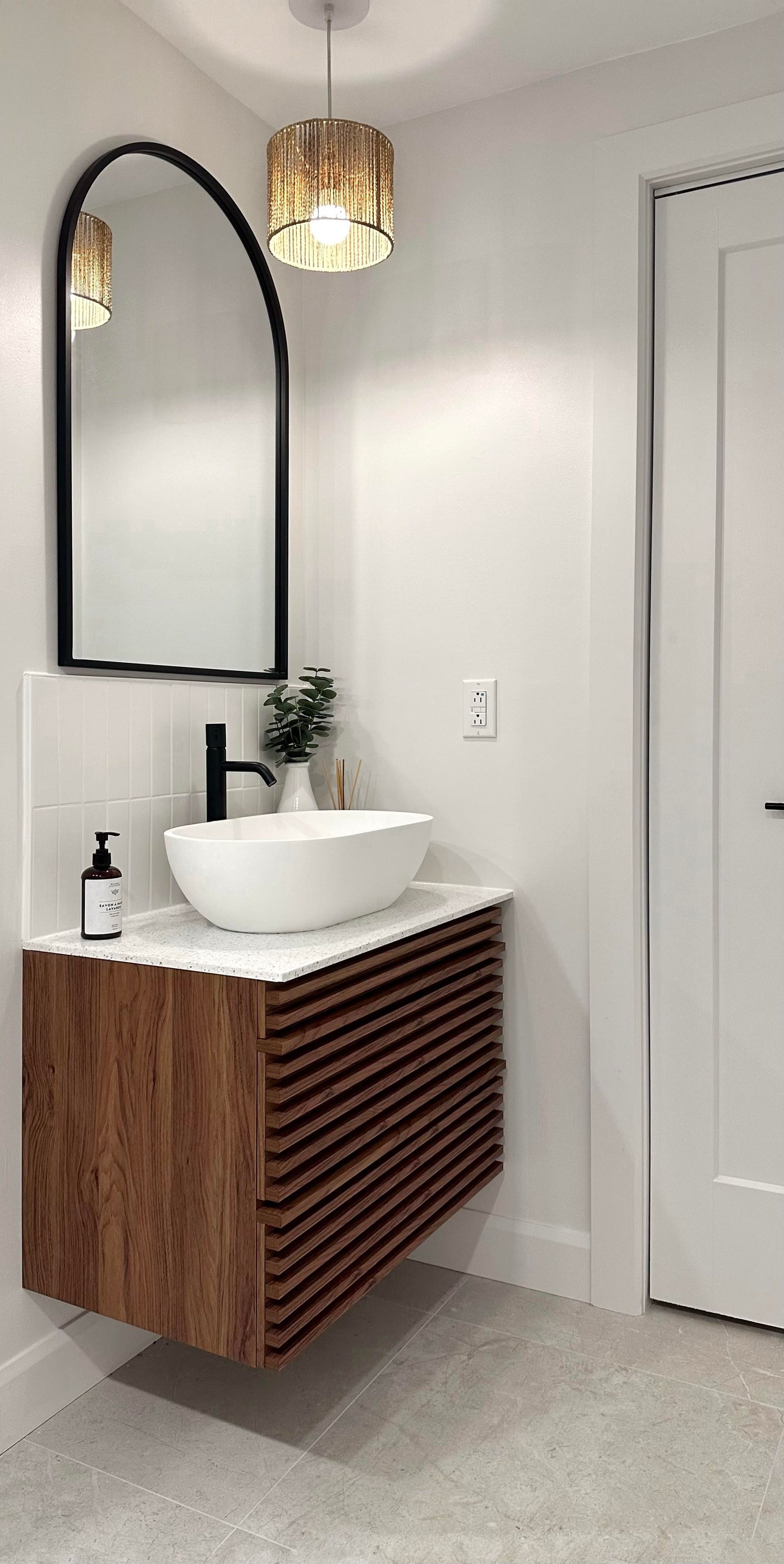 Salle de bains avec meuble-lavabo suspendu en bois, vasque blanche, miroir arqué et suspension lumineuse dorée.
