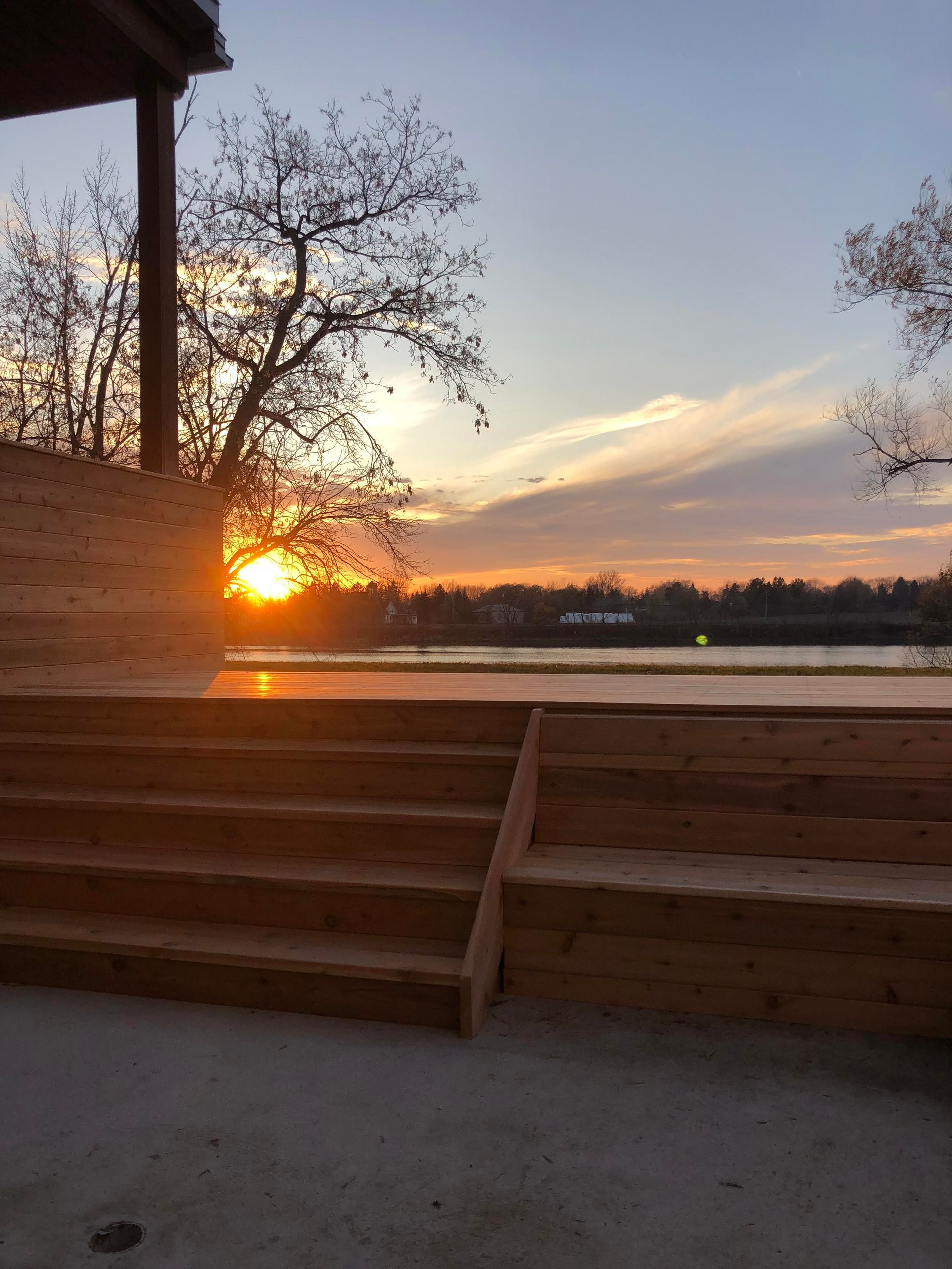 Des marches en bois mènent à une terrasse au bord de l'eau au coucher du soleil ; le soleil orangé brille derrière les arbres dénudés et les bâtiments au loin.