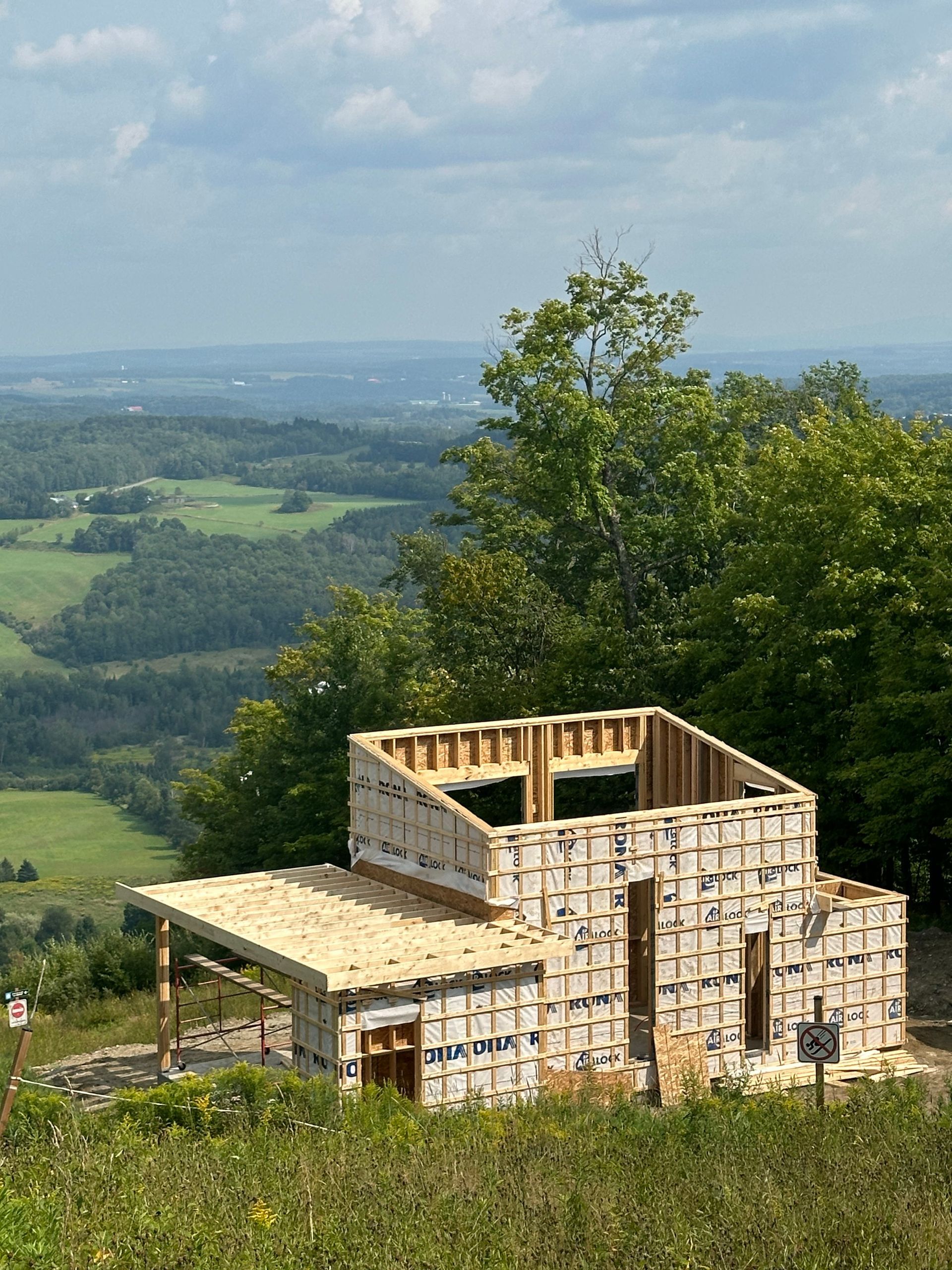 Maison en bois en construction au sommet d'une colline surplombant une vallée, des arbres et un paysage lointain.