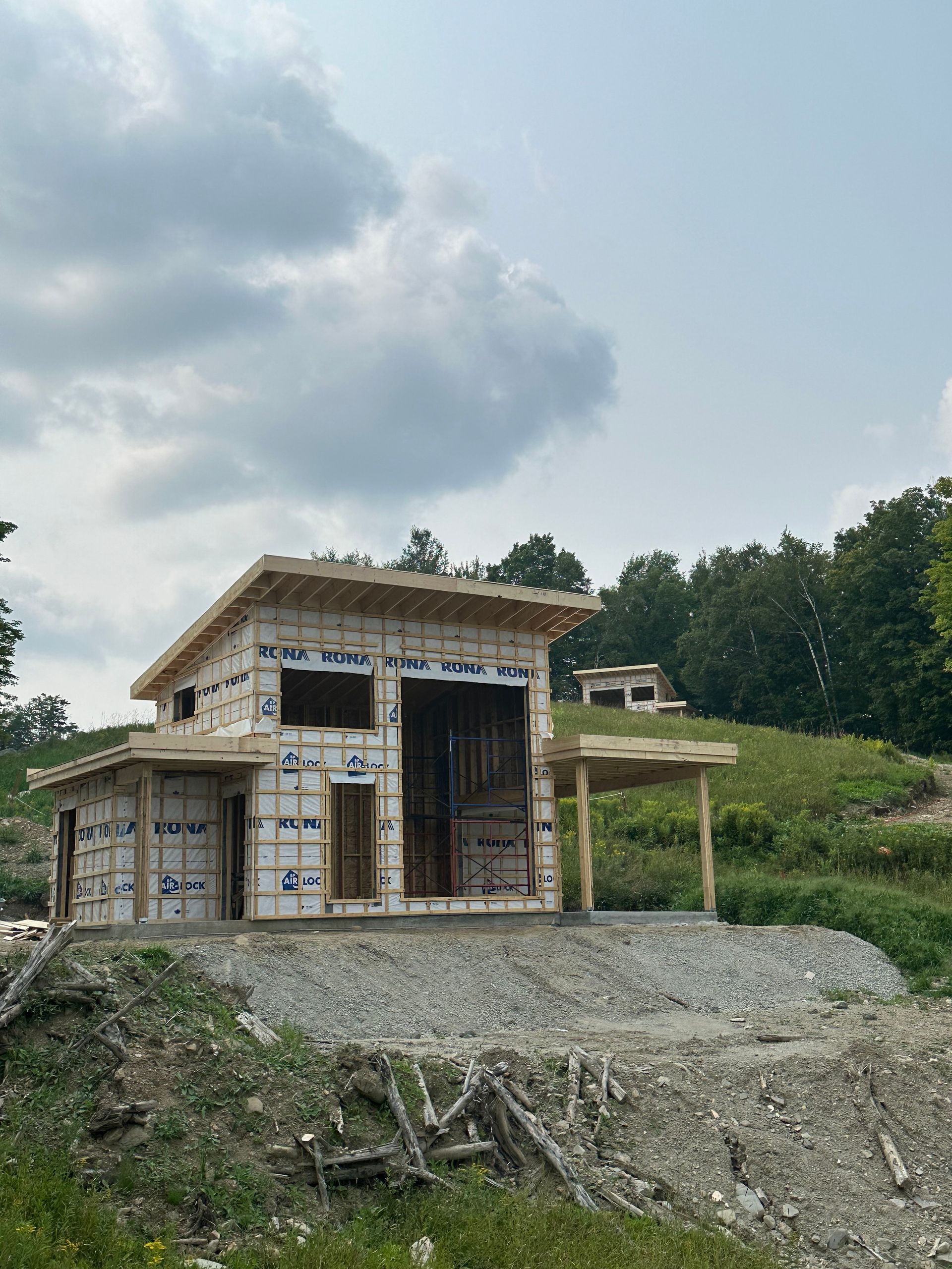 Chantier, petite maison inachevée à flanc de colline. Charpente extérieure visible, bâche bleue, toit vert. Ciel nuageux.