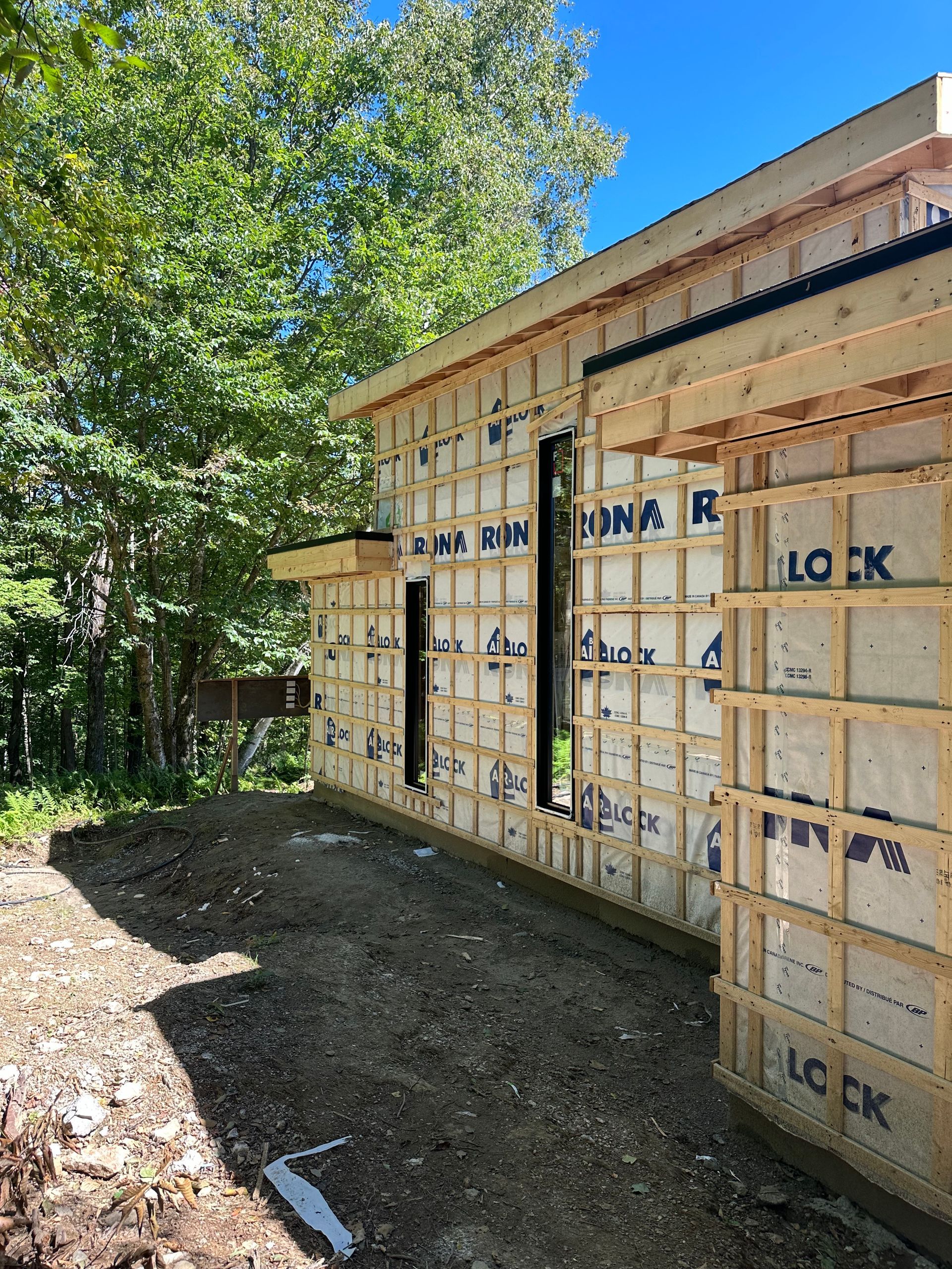 Extérieur d'un bâtiment en construction, montrant la charpente en bois, l'isolation et les ouvertures des fenêtres, à proximité d'arbres et d'un sol en terre battue.