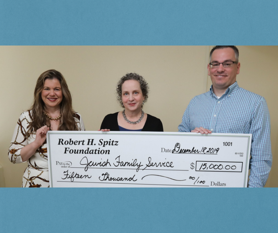 Three people hold a check for Jewish Family Service, donation for $15,000.  They are smiling in a room.