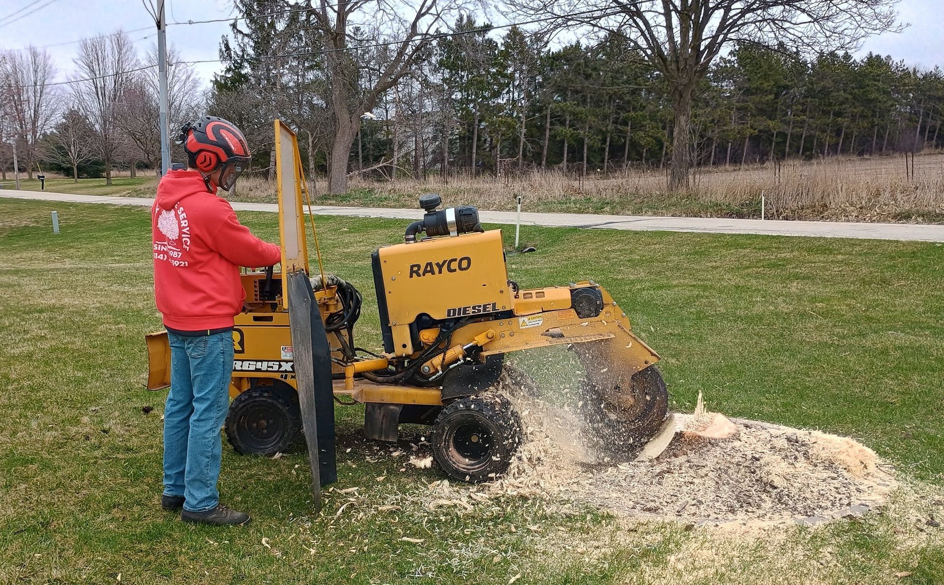 Person in red jacket operating a yellow wood chipper outdoors, spraying wood chips onto grass.