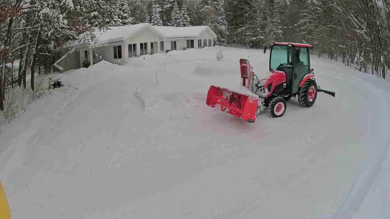 Red tractor plowing snow in a driveway near a house and trees on a snowy hillside